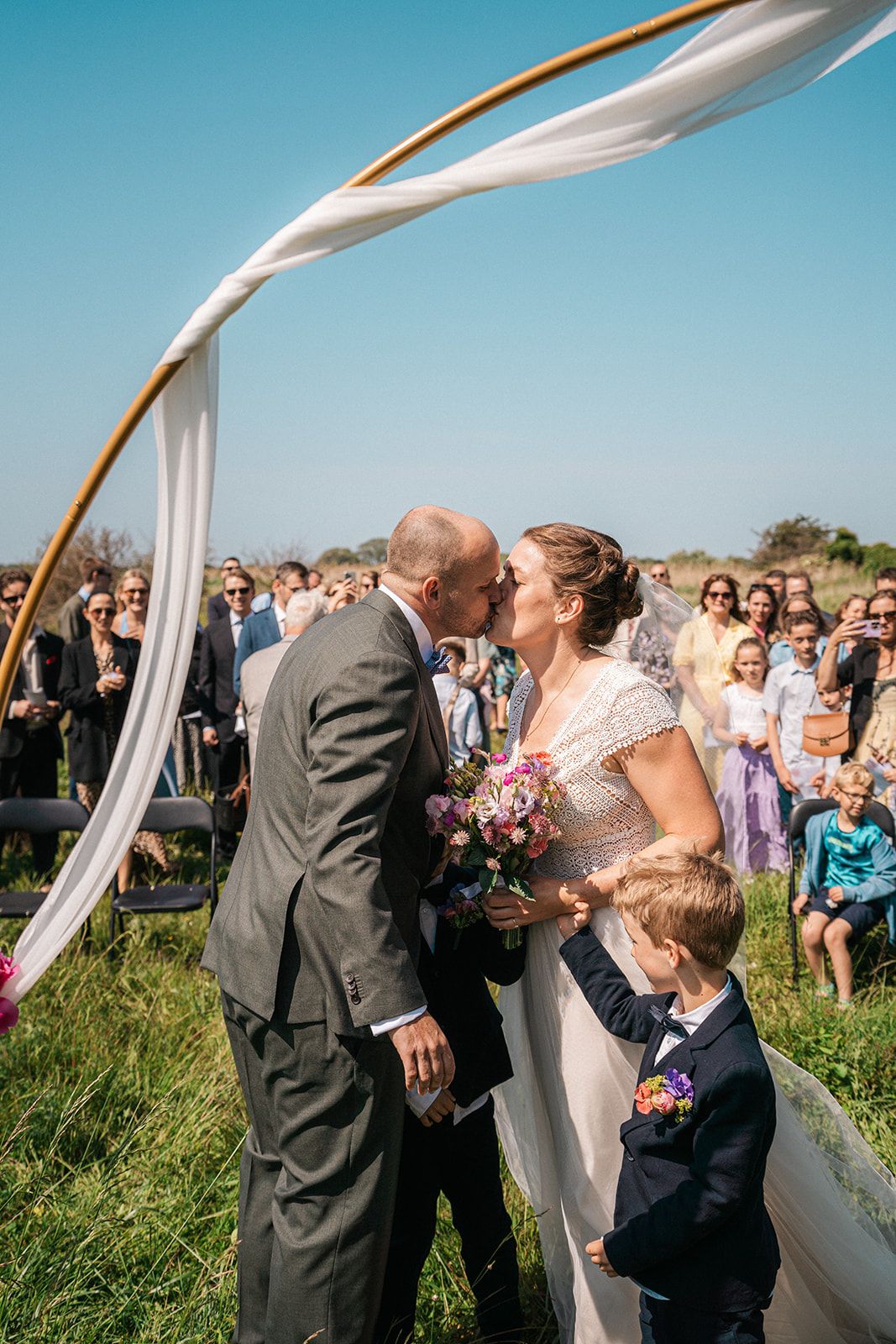 An outdoor wedding ceremony under a white fabric canopy with guests watching.