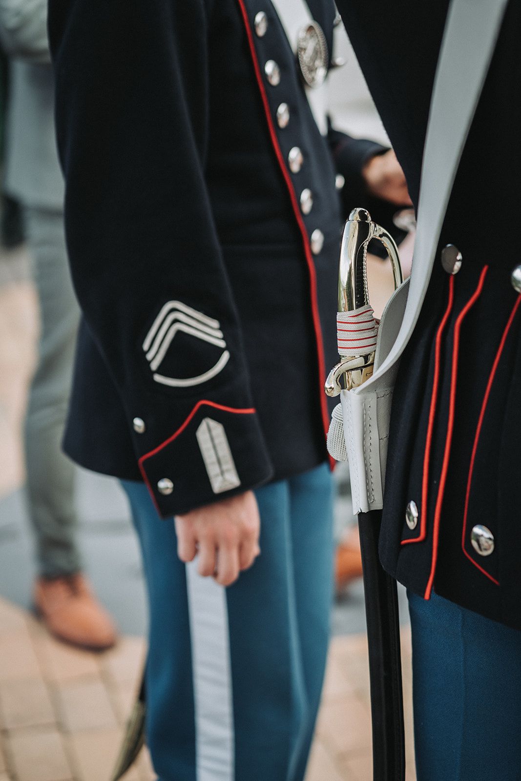 Close-up detail of military dress uniform with black jacket featuring red piping, silver buttons and ceremonial sword.