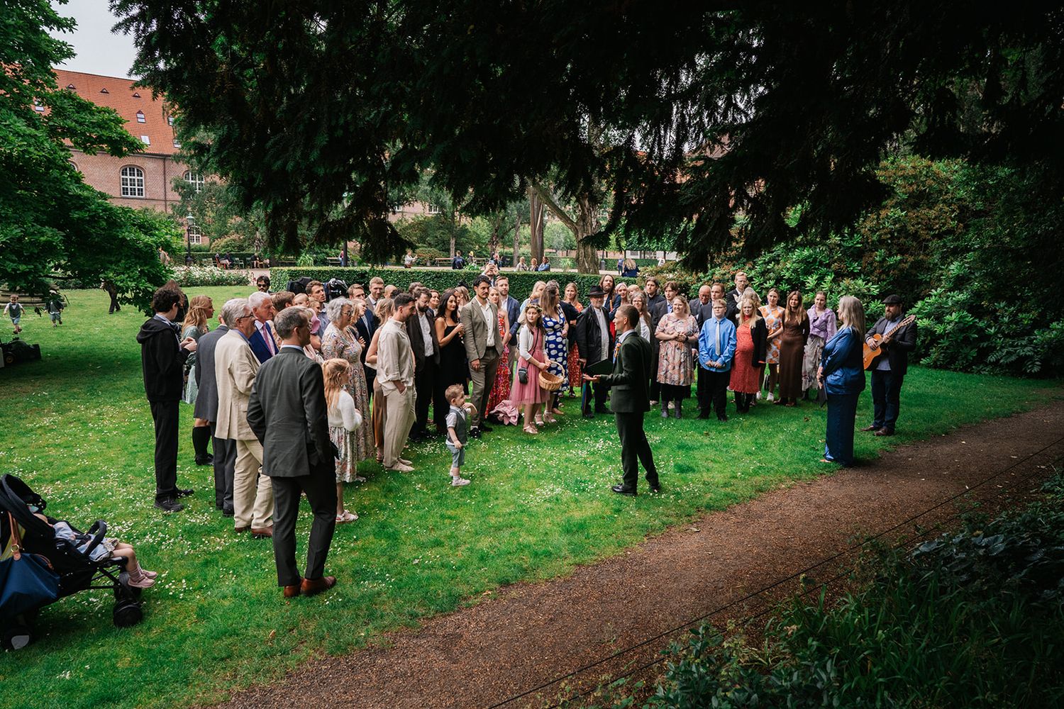 Wedding guests gather in a tranquil garden setting as they walk along a tree-lined dirt path on a sunny afternoon.