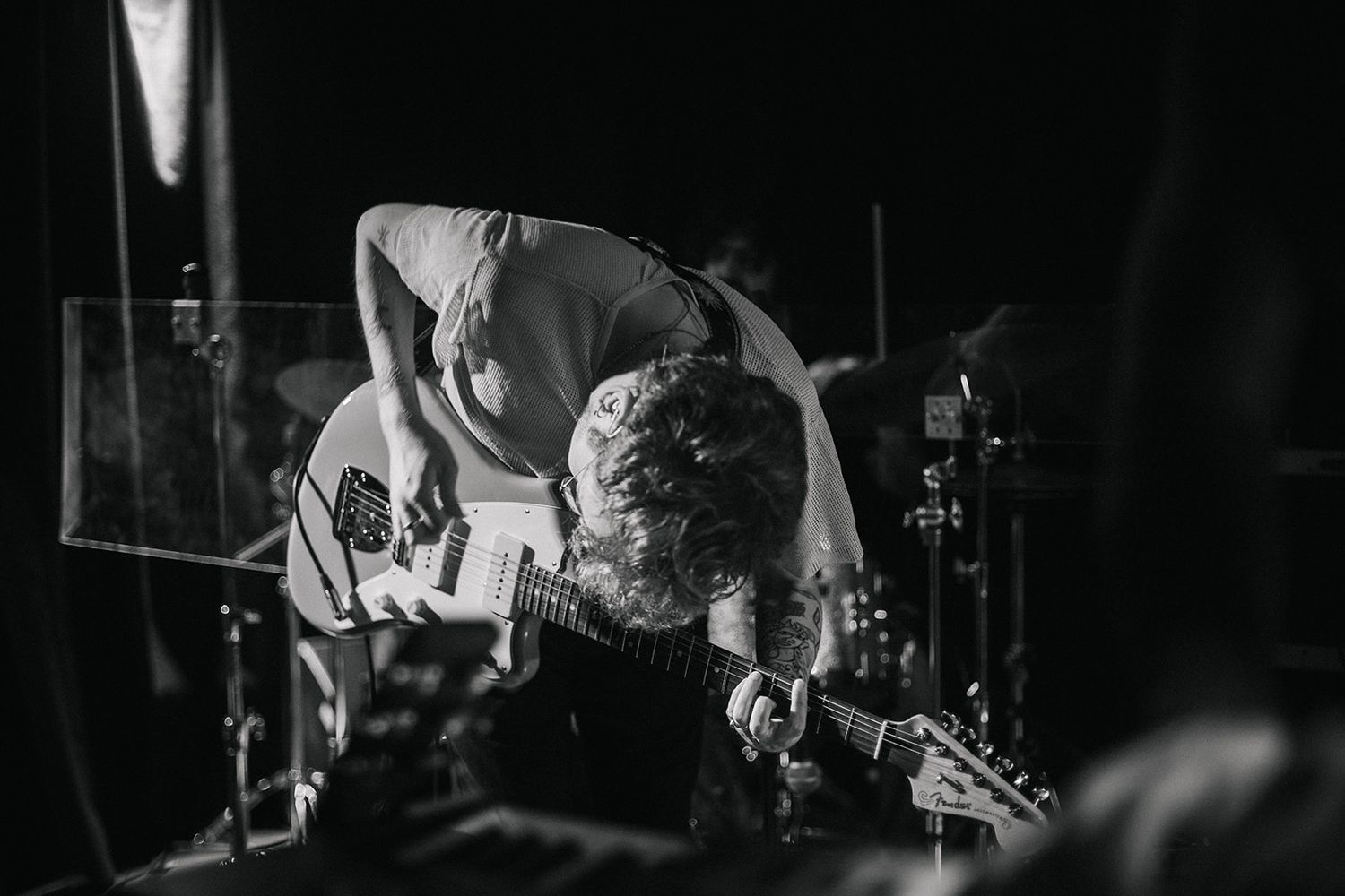 Black and white photo of a performer bending over while playing electric guitar on a dark stage during a live show.