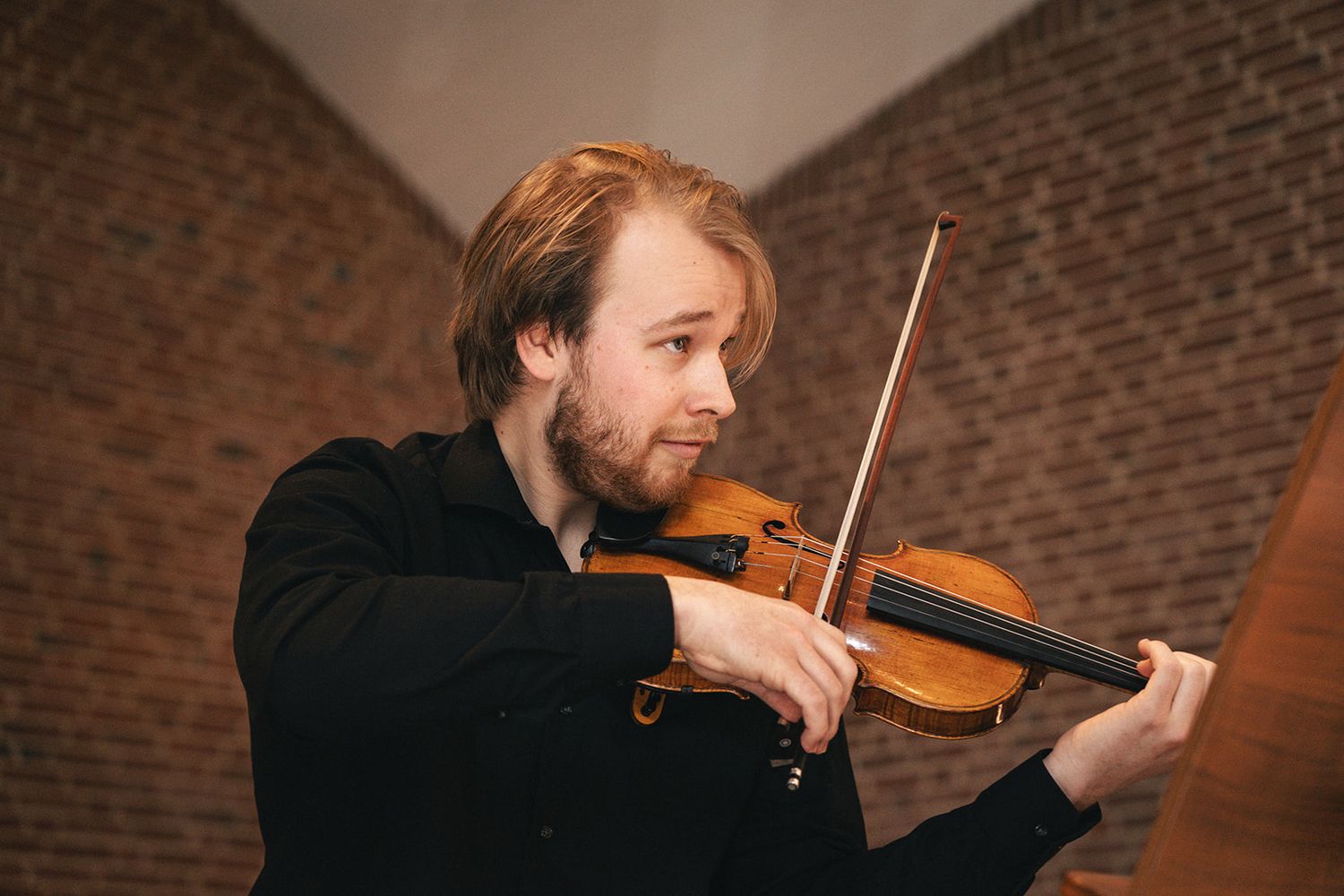 A musician plays violin with passion against a rustic brick wall background in dimmed lighting.