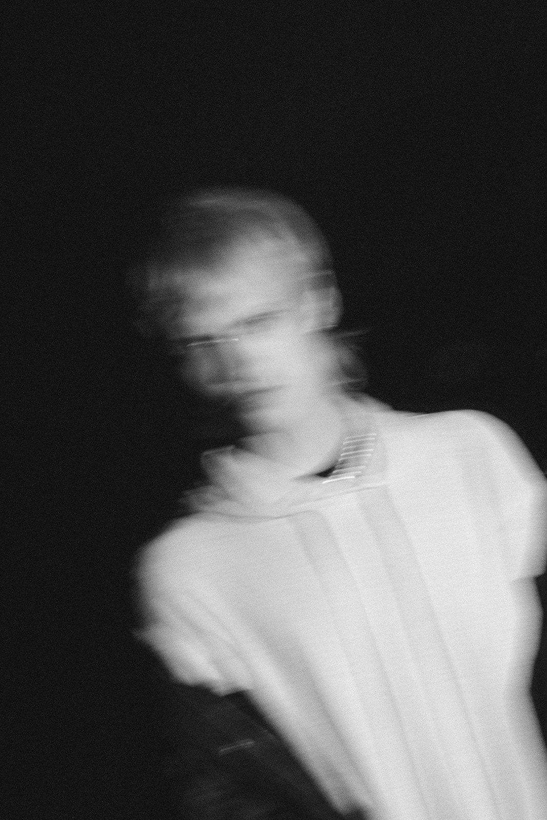 Black and white motion blur portrait with blurred movement effect against dark background showing figure in white clothing.