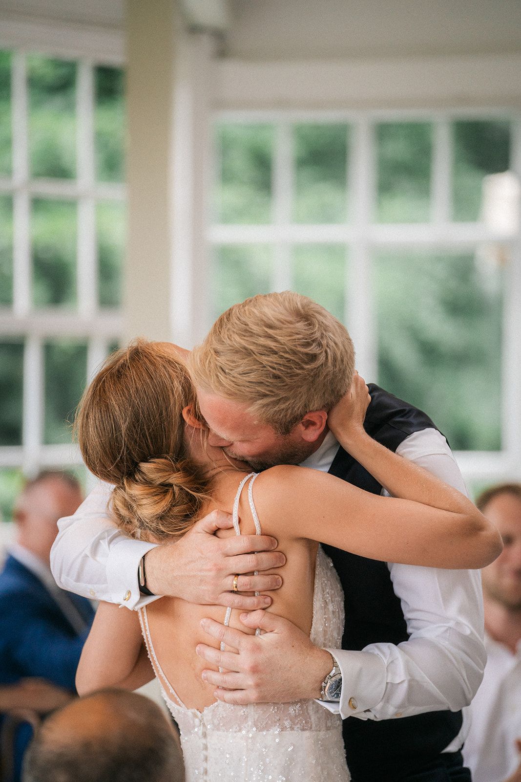 A romantic kiss between newlyweds sharing their first dance at a wedding reception with sunlit windows in the background.