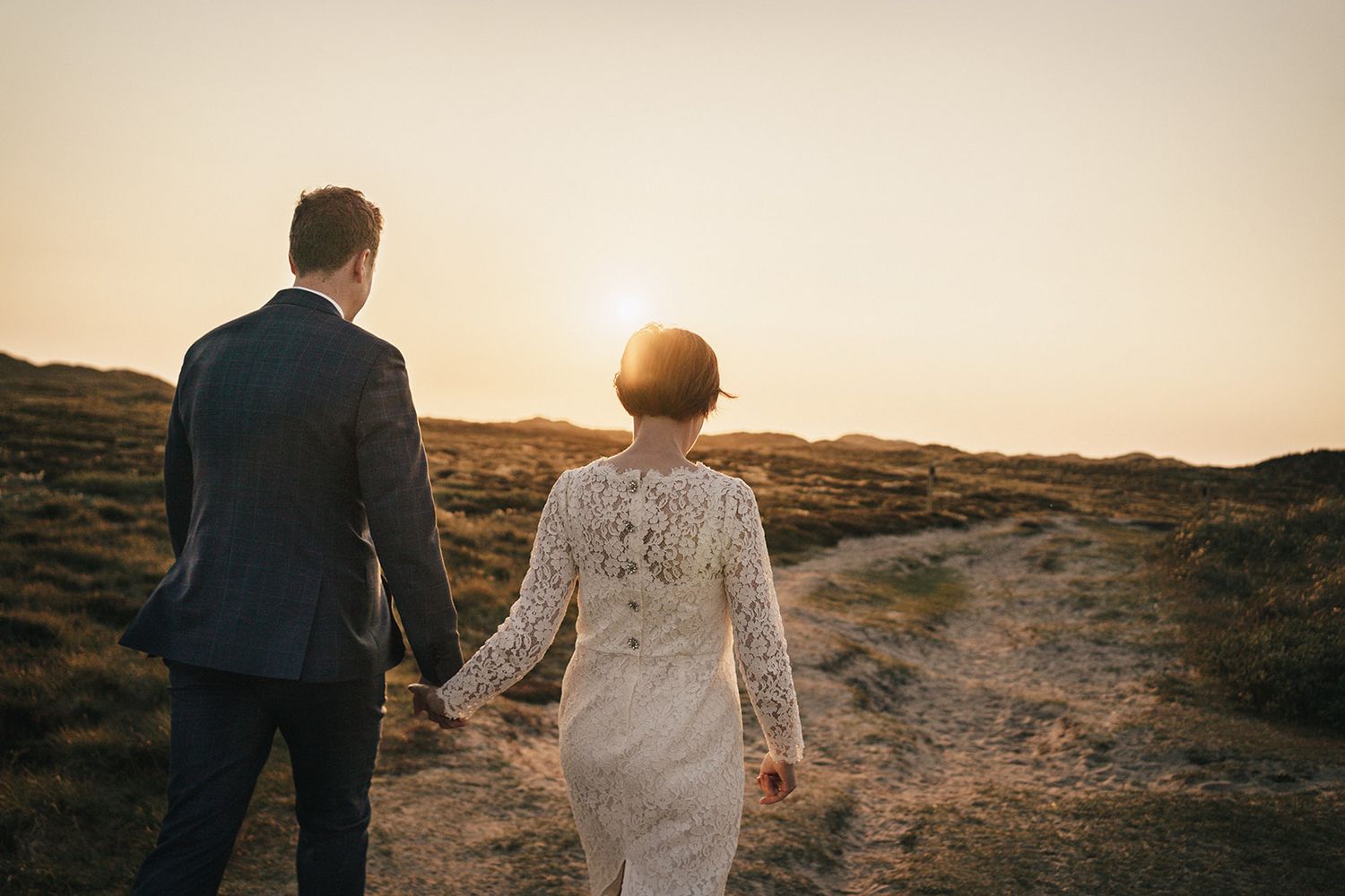 A couple in wedding attire walks hand in hand during golden hour sunset on a rugged hillside path.