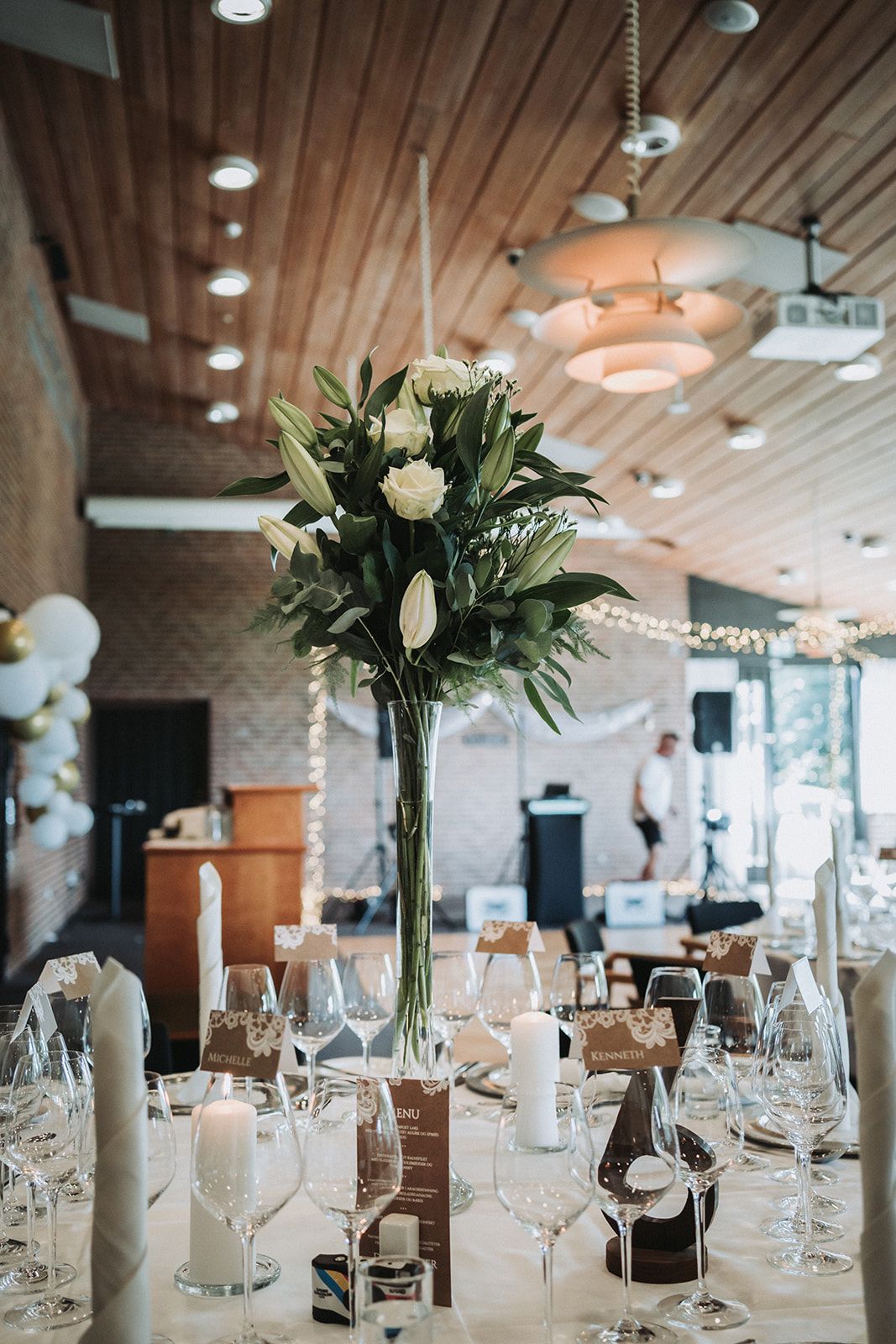 Elegant wedding reception table with tall white lily centerpiece and wine glasses in modern venue with wood ceiling.