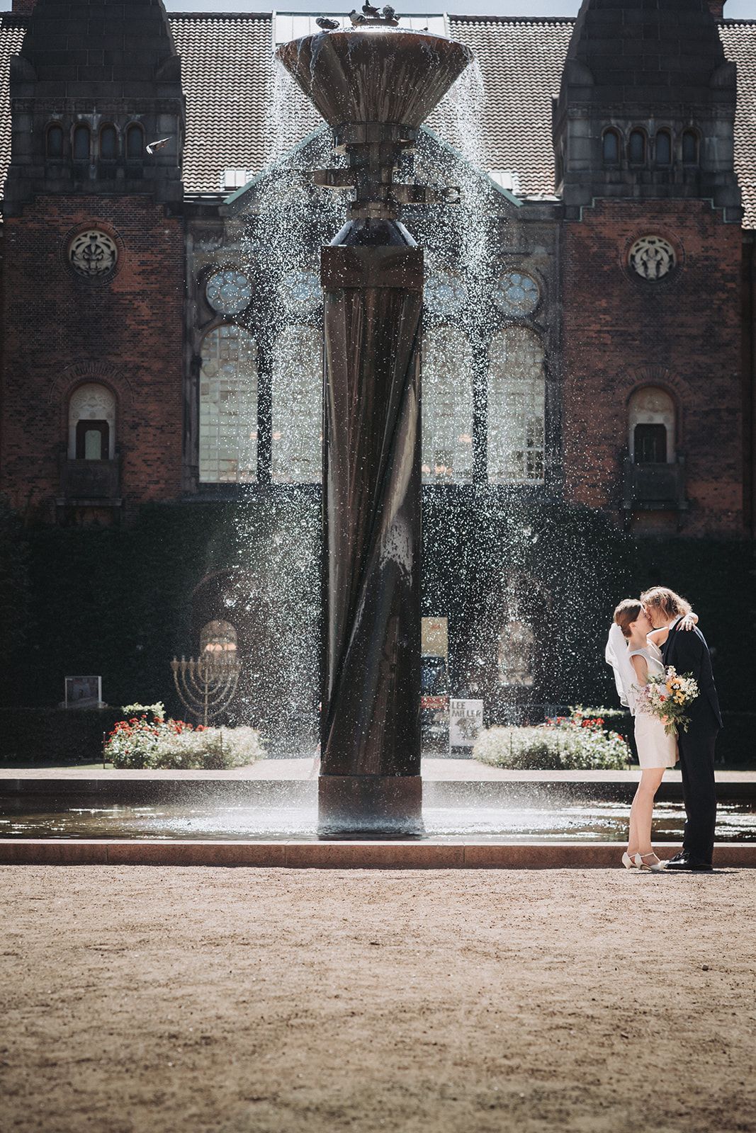 Modern twisted fountain sculpture sprays water in front of historic brick building with romantic couple silhouetted nearby.
