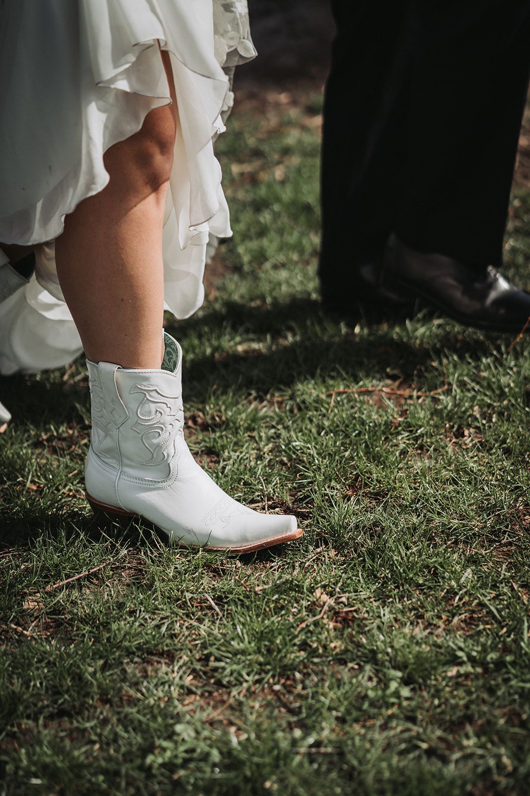 White cowboy boots peek out beneath a wedding dress on grassy ground next to formal black shoes at an outdoor ceremony.