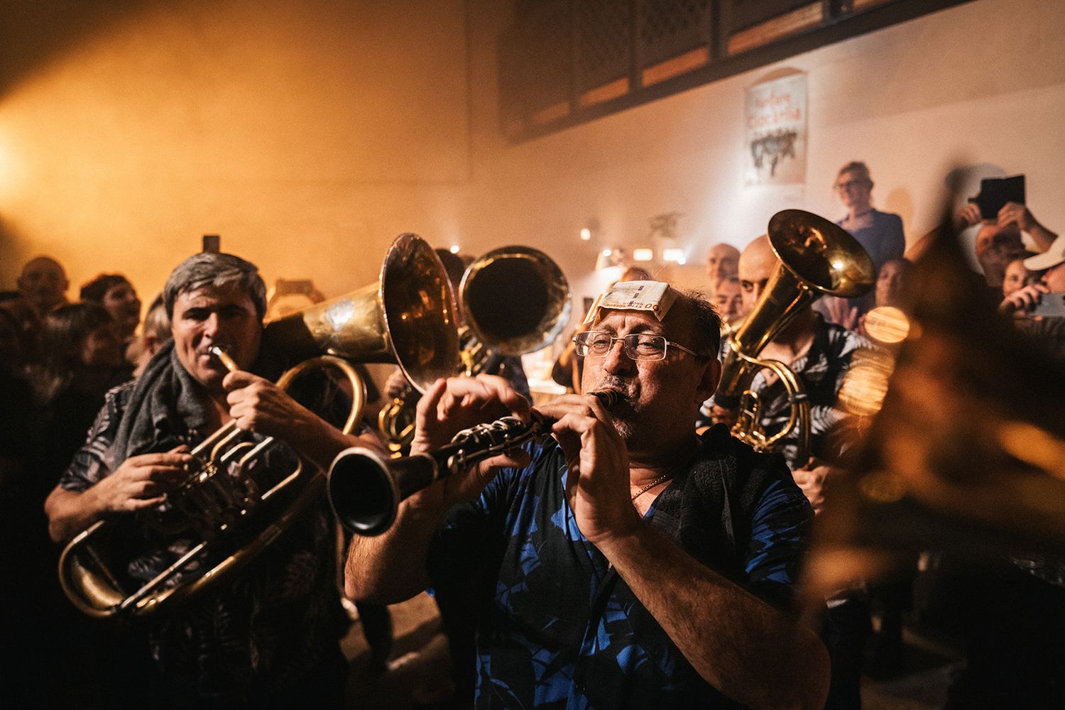 Brass horn musicians perform together in a dimly lit venue with warm lighting creating an atmospheric scene.