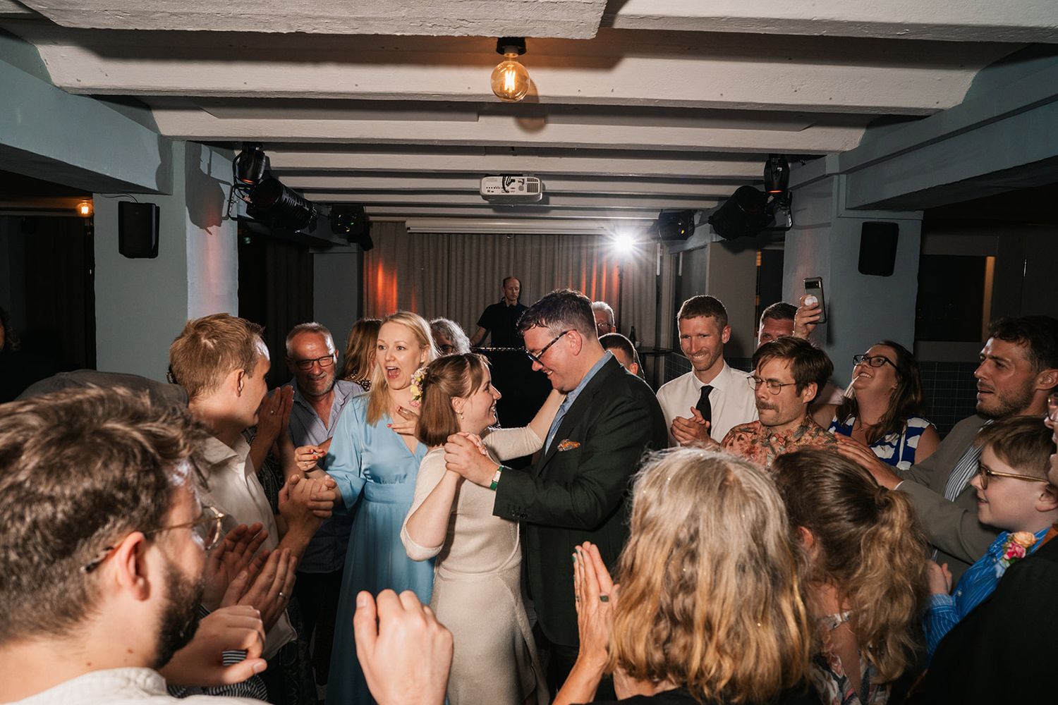 A crowded dance floor at a wedding reception with guests dancing and celebrating under wooden ceiling beams.