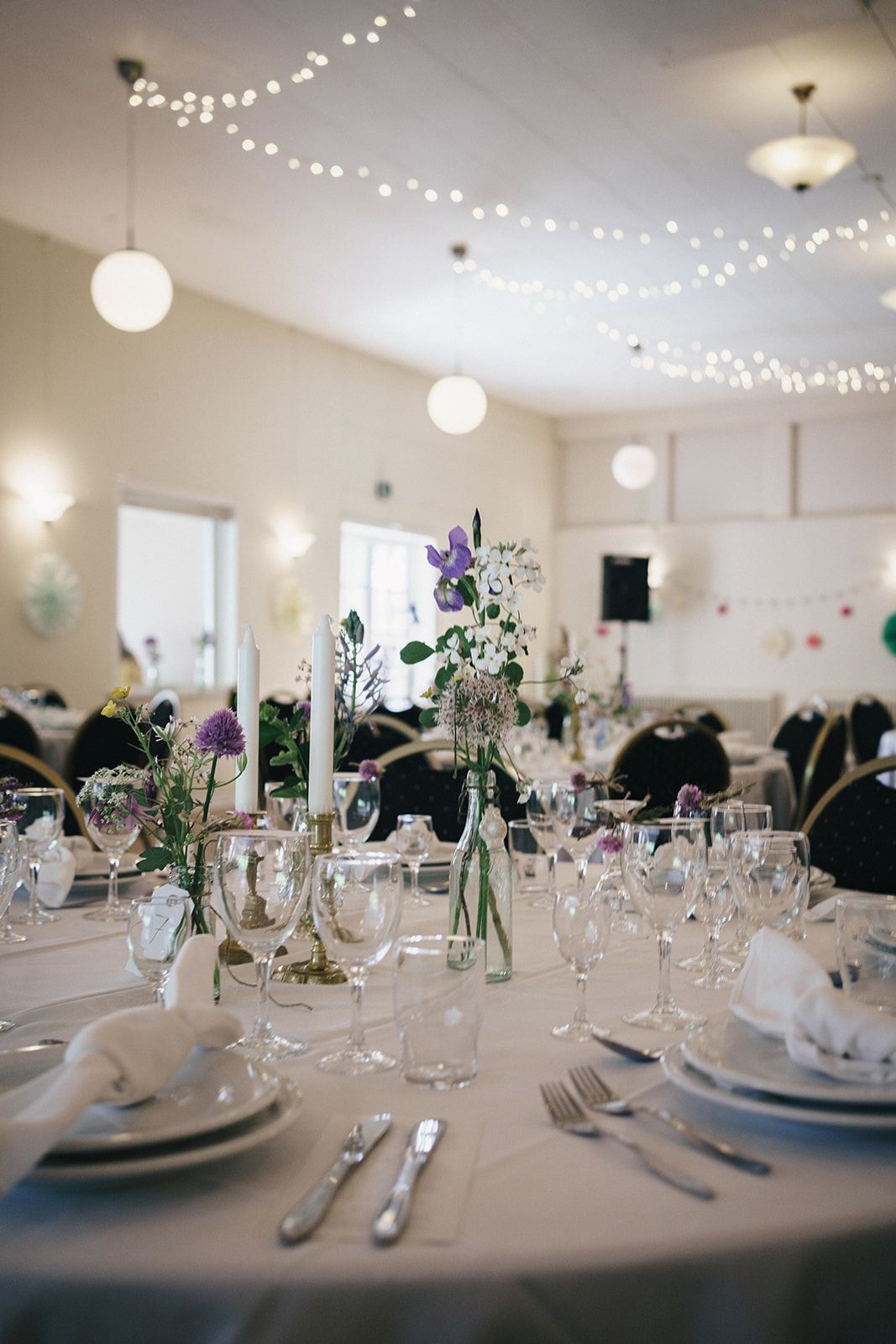 Elegant wedding reception table setting with purple flowers, string lights, and white paper lanterns overhead.