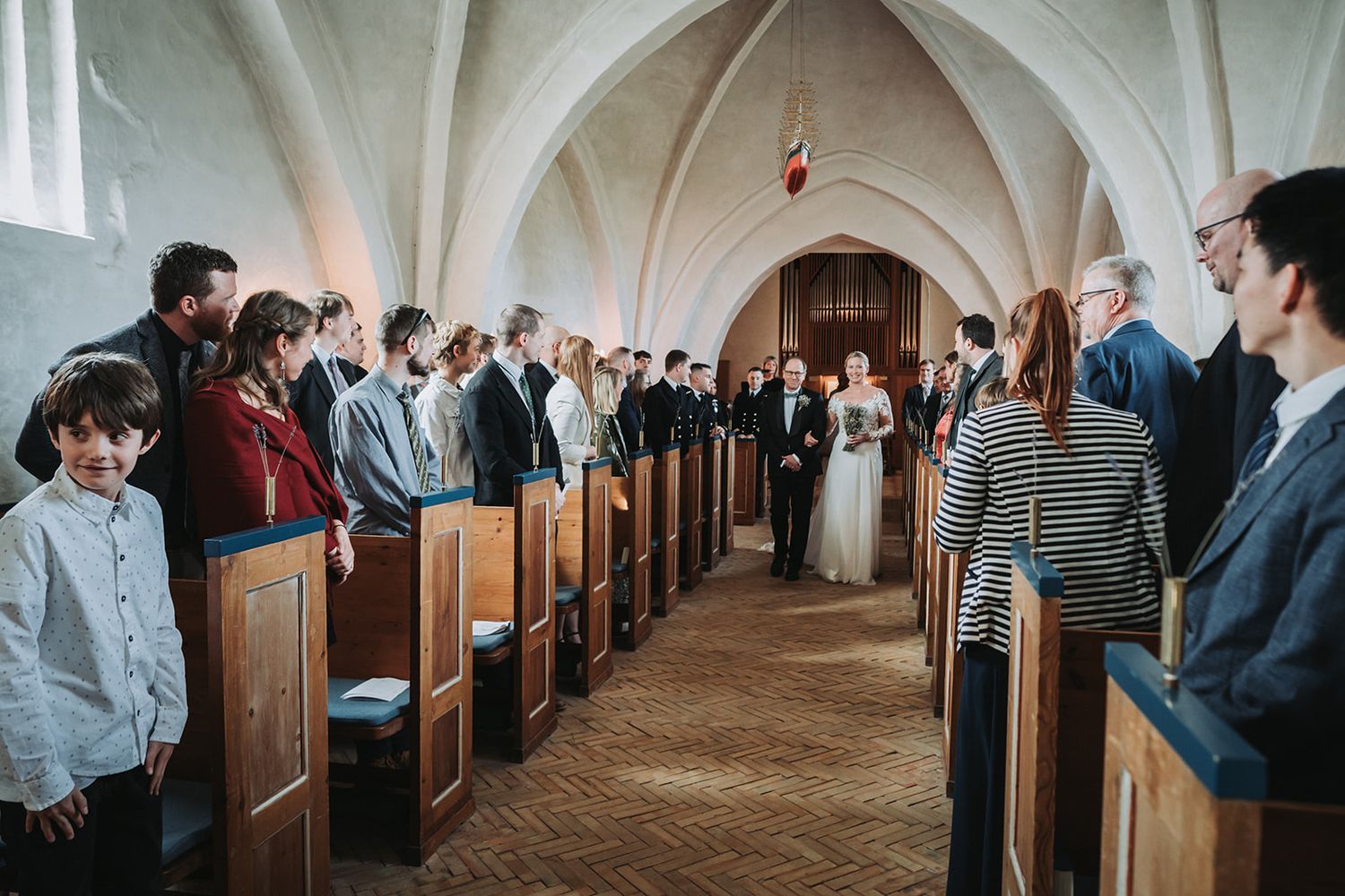 Mourners sit solemnly in wooden pews during a church service inside a bright white vaulted sanctuary.