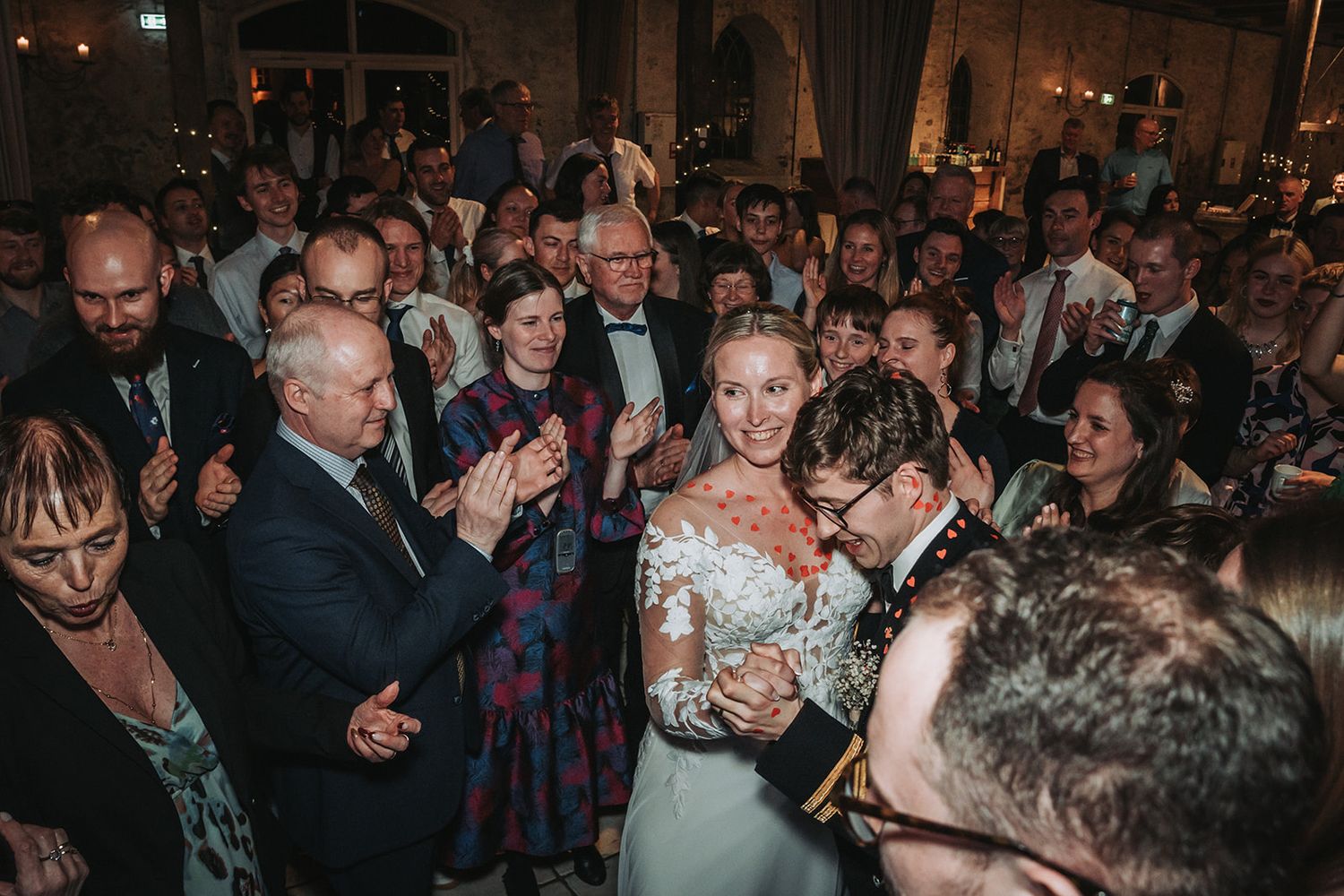 Wedding reception guests gather on a crowded dance floor celebrating with the newlyweds during an evening celebration.