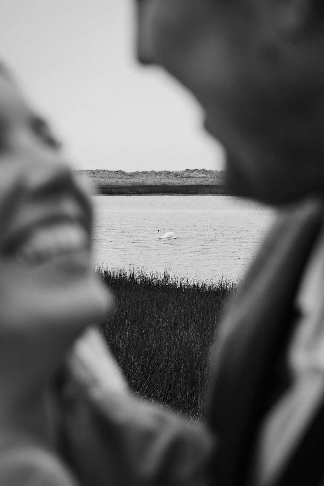 Black and white coastal scene with marsh grass and water in the background framed by blurred figures in foreground.