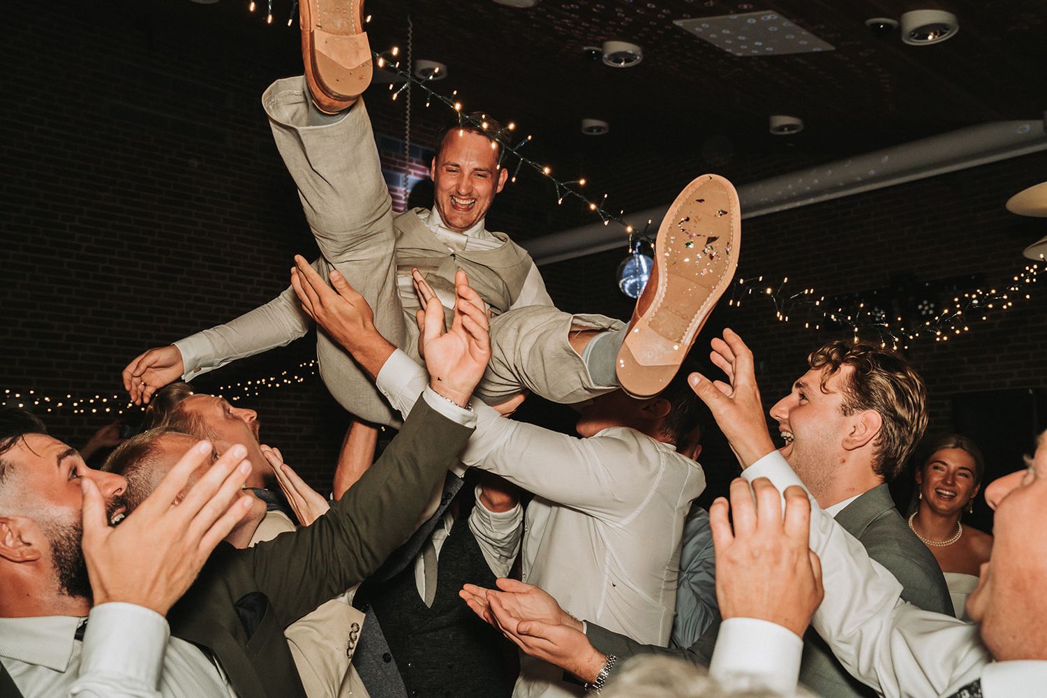 Crowd of wedding guests in grey suits lift and carry someone during an energetic reception celebration dance party.