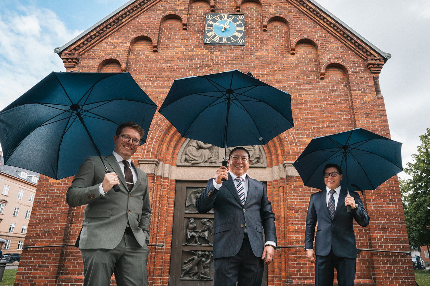 Three well-dressed figures stand outside a brick church building holding dark blue umbrellas on an overcast day.