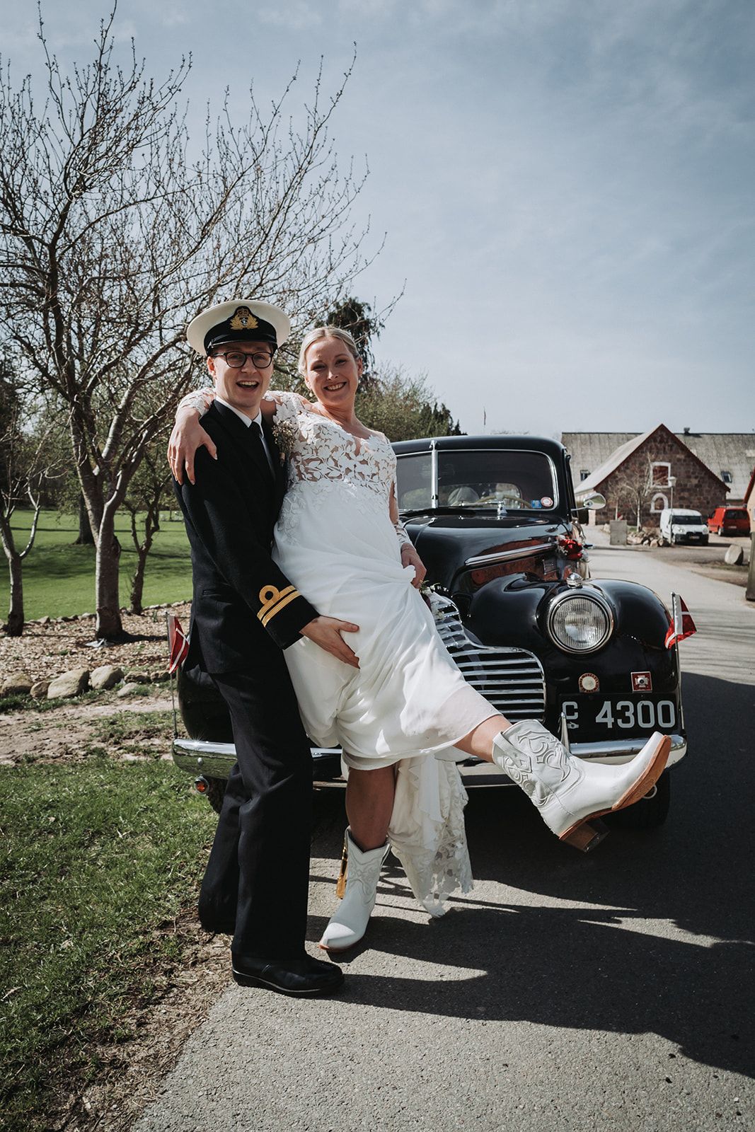 A vintage black car with license plate P4300 parked near a couple in wedding attire posing for a photo.