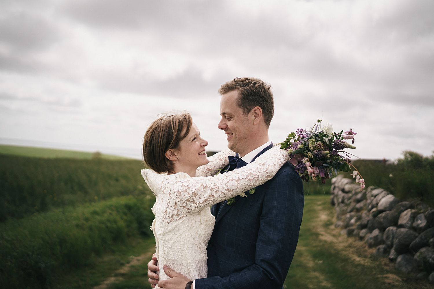 A couple embraces in a romantic moment against a cloudy countryside backdrop with stone walls and rolling hills.