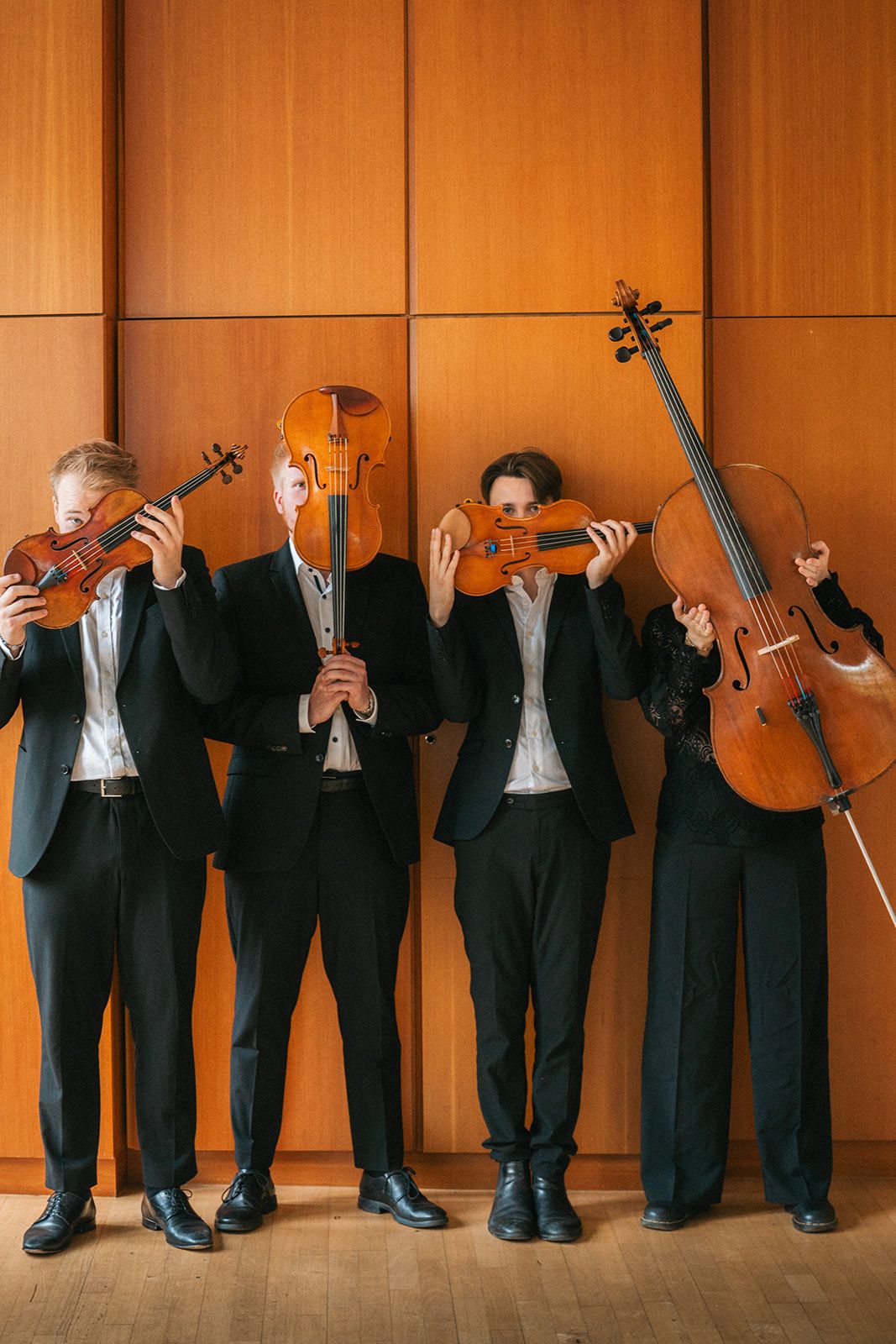 String quartet members playfully hide their faces behind their instruments against orange wall.