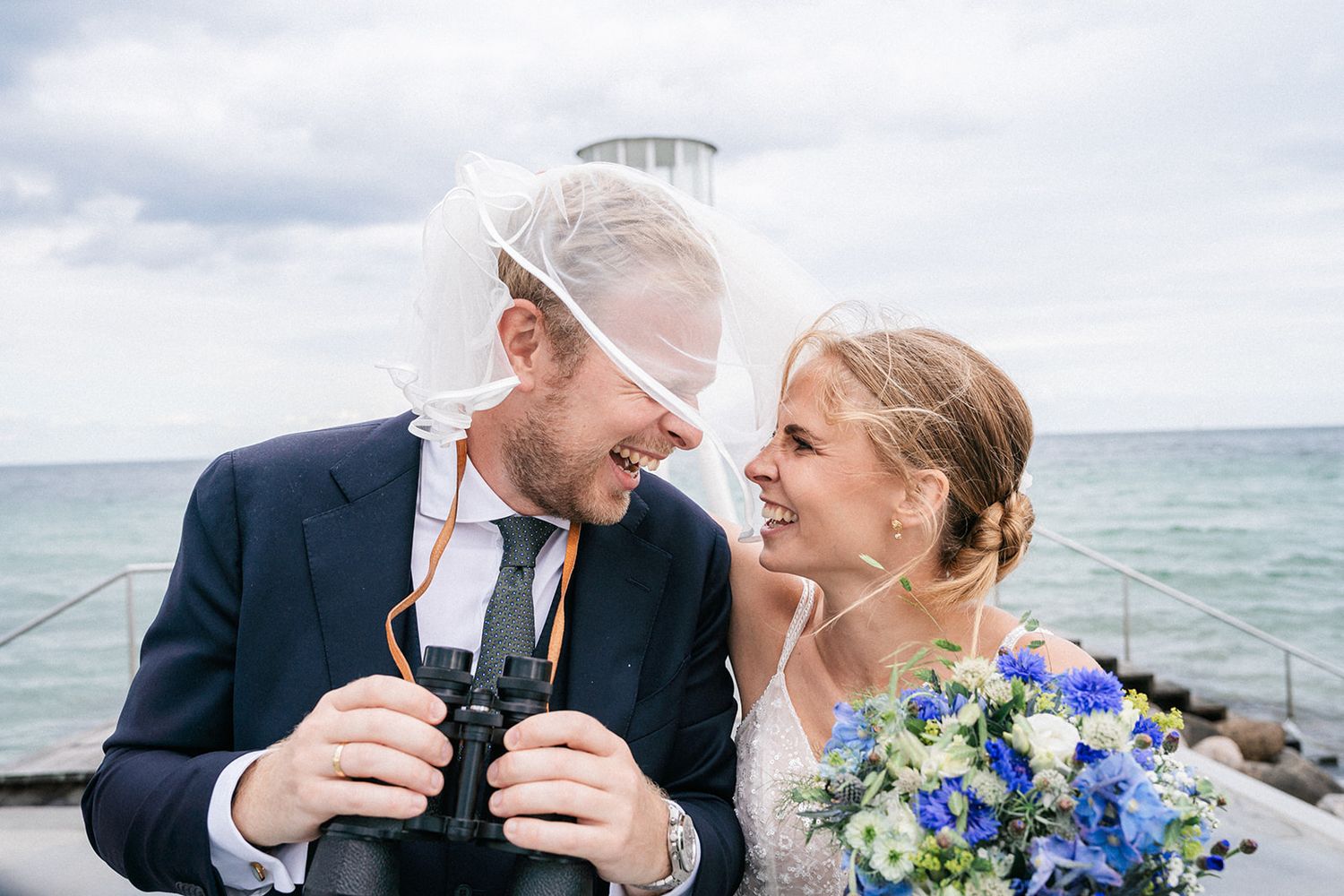 Happy newlyweds share a laugh while holding champagne glasses on a coastal overlook with blue flowers in the bouquet.