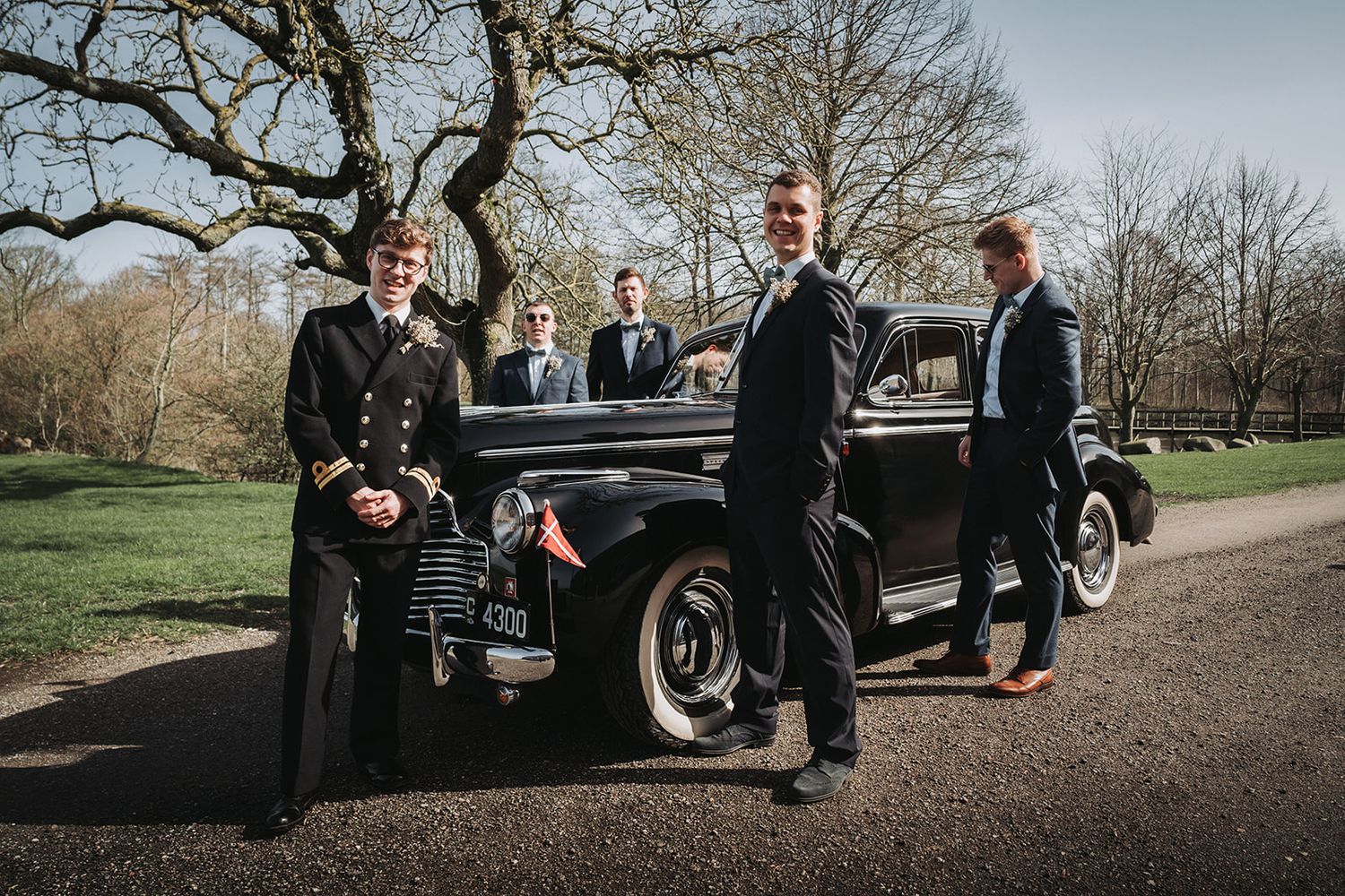A vintage black car surrounded by people during a wedding celebration with bare trees in the background.