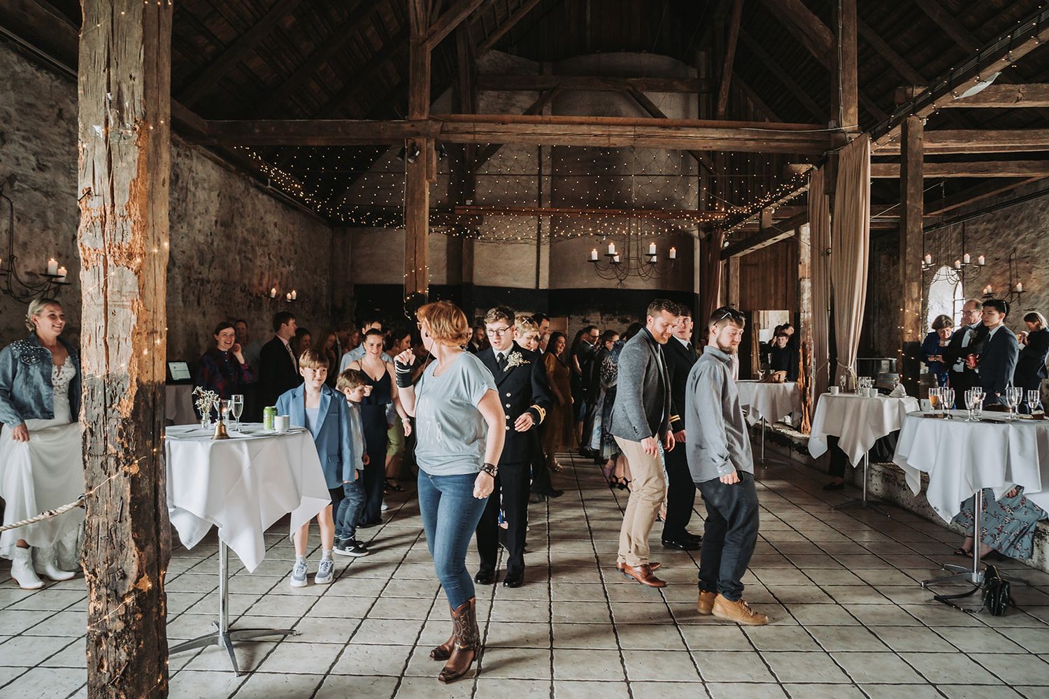 Guests mingle around white-clothed tables at a rustic barn wedding reception with exposed wooden beams and vintage charm.