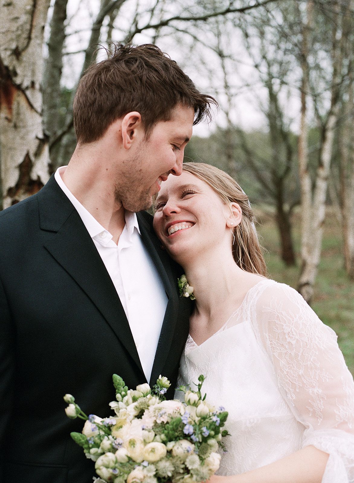 A romantic couple shares tender moments in a wooded setting while holding a delicate white and green bouquet.