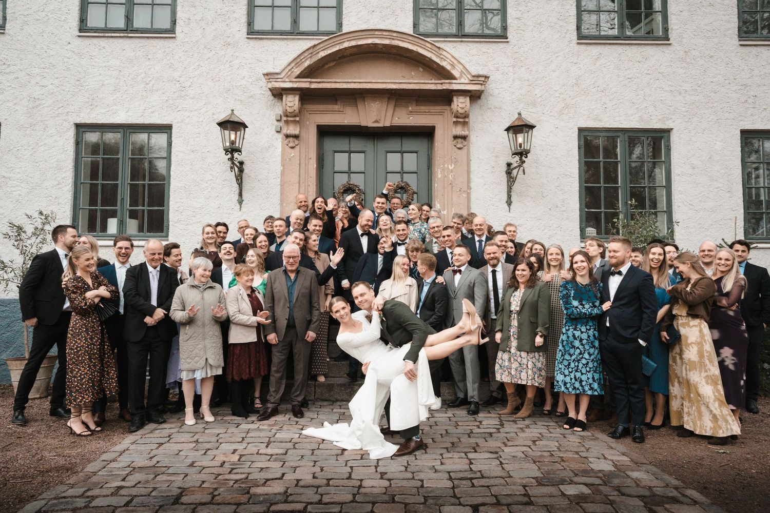 Large wedding group photo outside a historic manor, bride being lifted by groom surrounded by elegantly dressed guests.