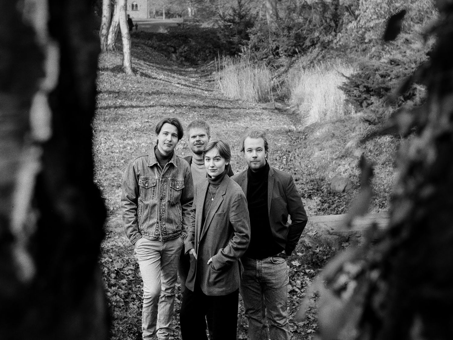 Three people in dark jackets stand together outside on a wooded path during autumn.