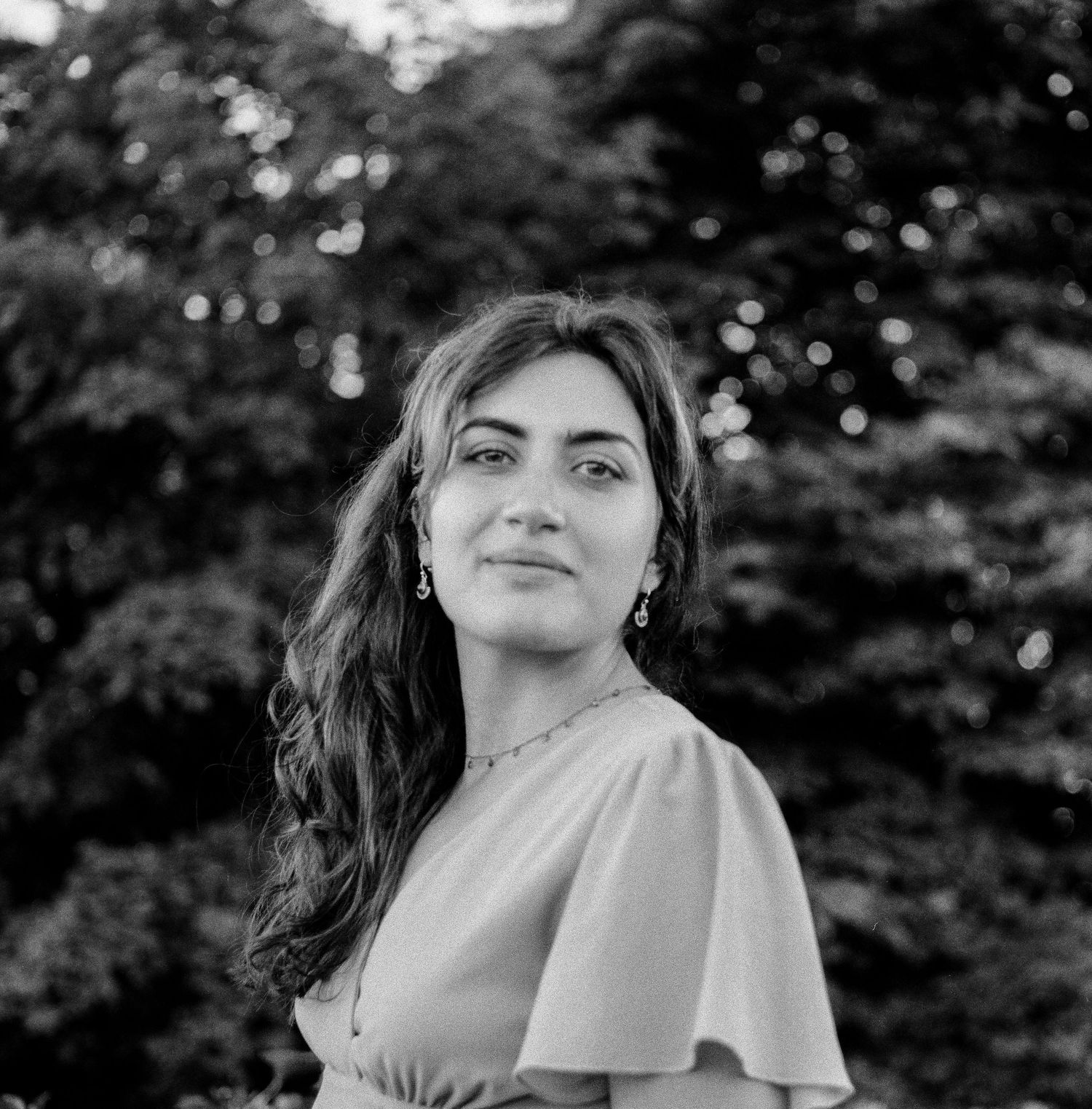 Black and white portrait of a person with long dark hair and earrings smiling outdoors with trees in background.