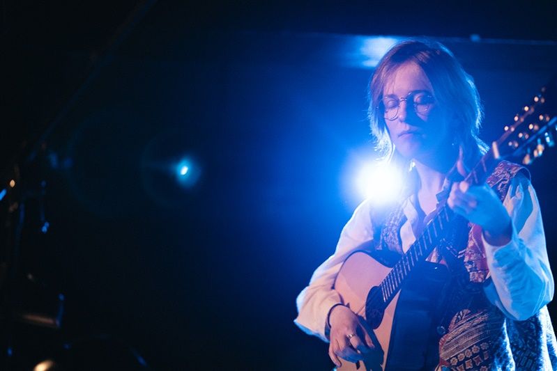 Musician performs on acoustic guitar under dramatic blue stage lighting while bathed in bright spotlight.