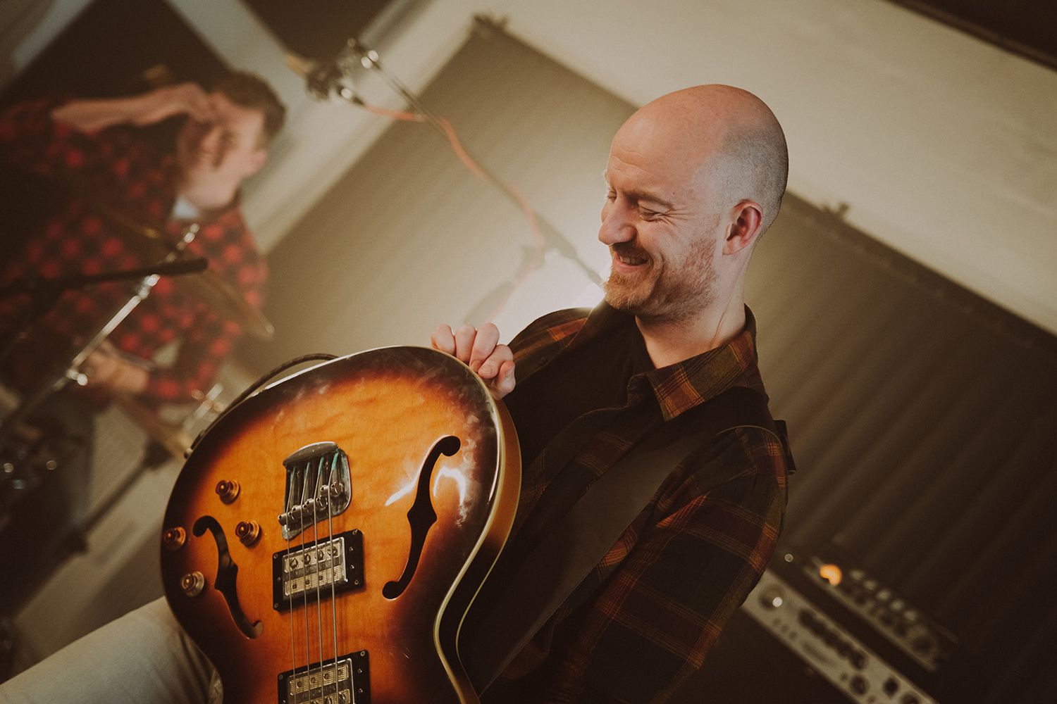 Musician smiles while holding warm-toned hollow body electric bass guitar in recording studio setting.