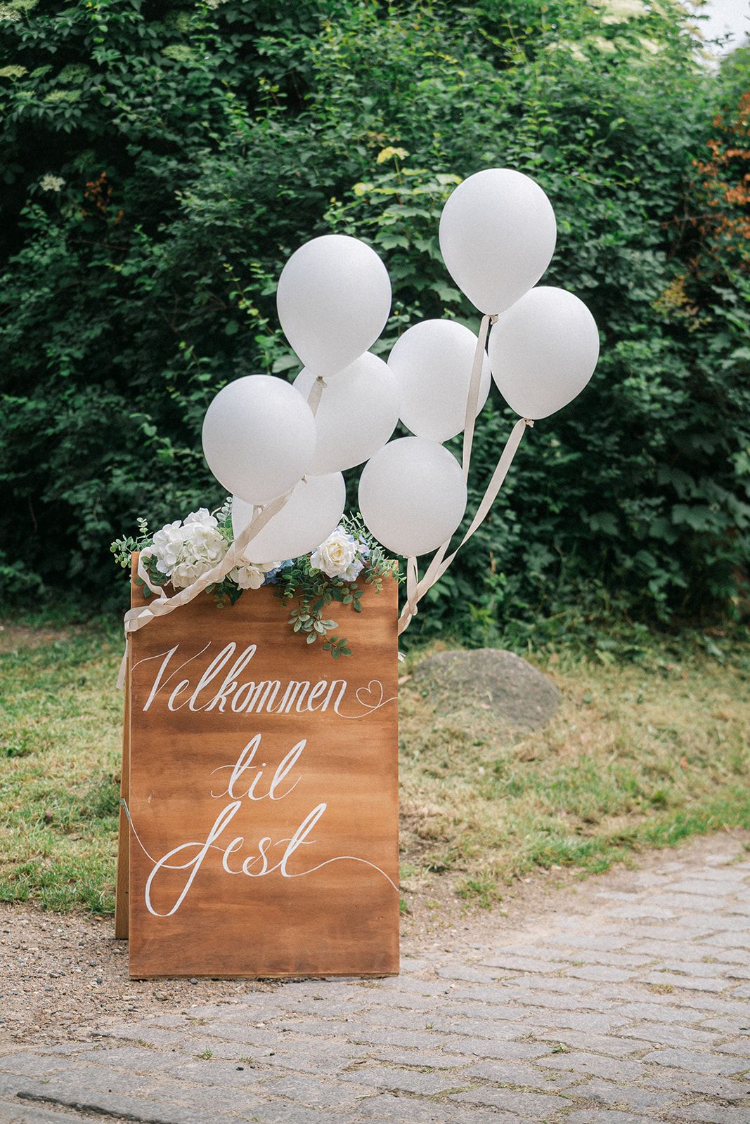 Wooden welcome sign with white calligraphy and white balloons against green foliage background.