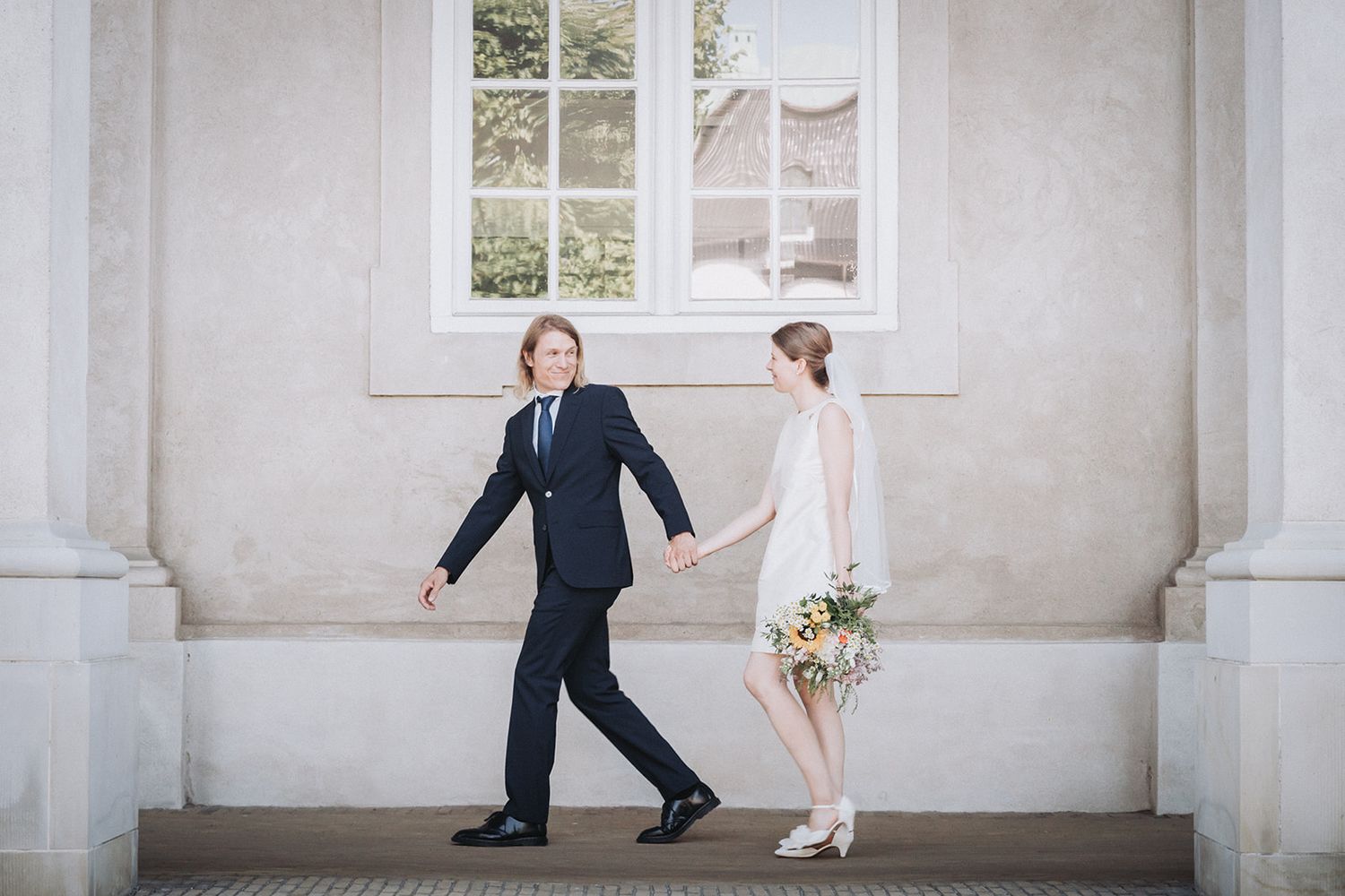 Bride and groom share a romantic embrace while walking along a grand white stone colonnade.