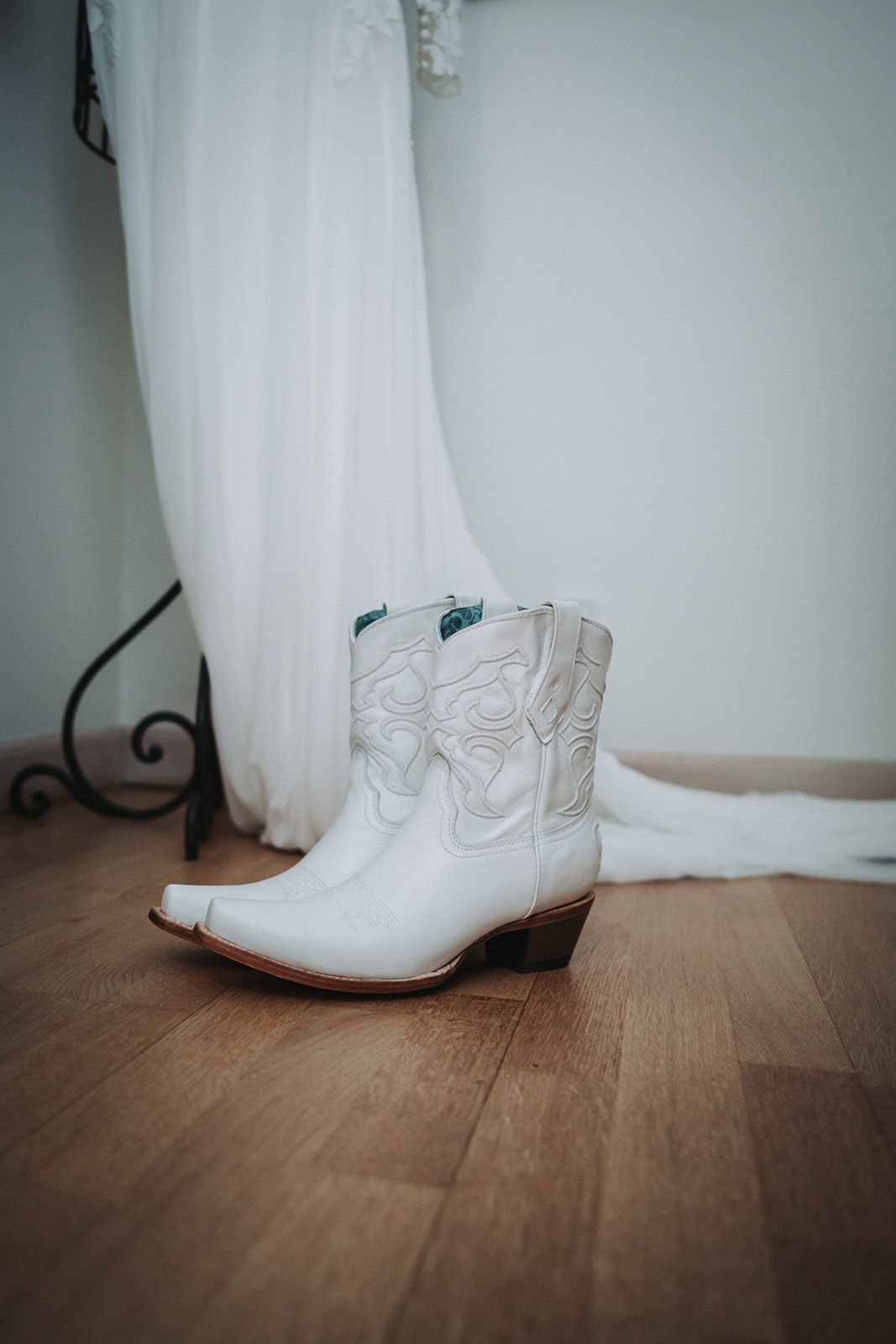 White wedding cowboy boots sitting on a hardwood floor next to flowing white fabric.
