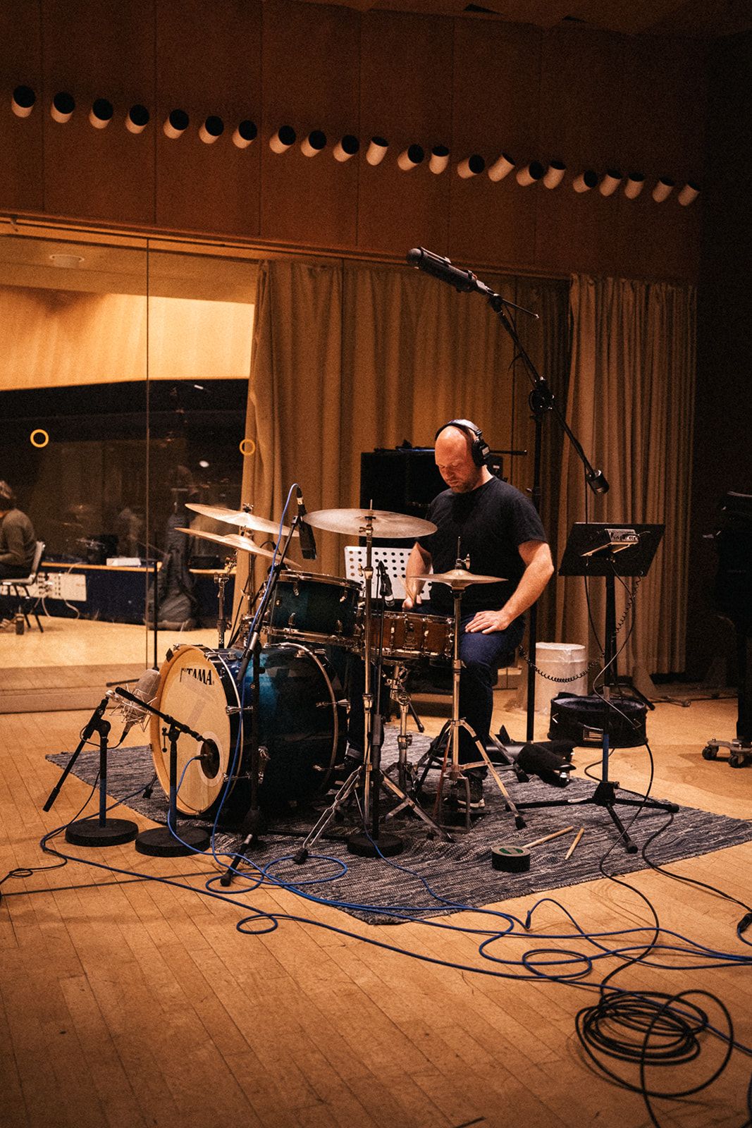 A drummer performs in a recording studio with microphones and equipment set up on a wooden floor.