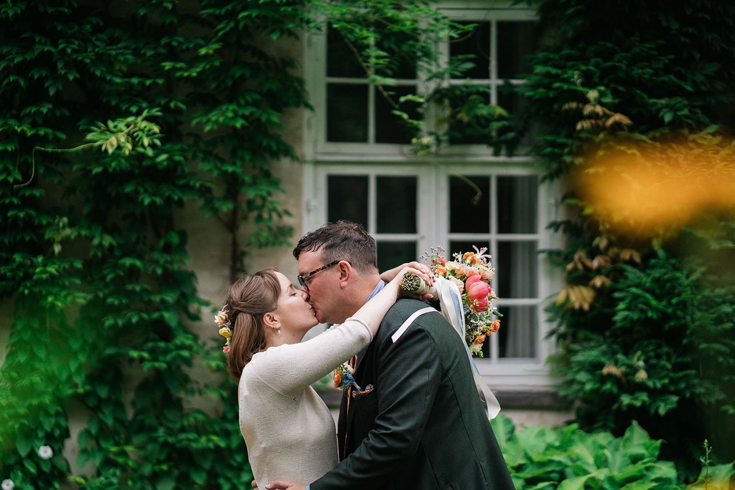 Wedding couple shares a romantic kiss outdoors in a lush green garden setting with white window frame backdrop.