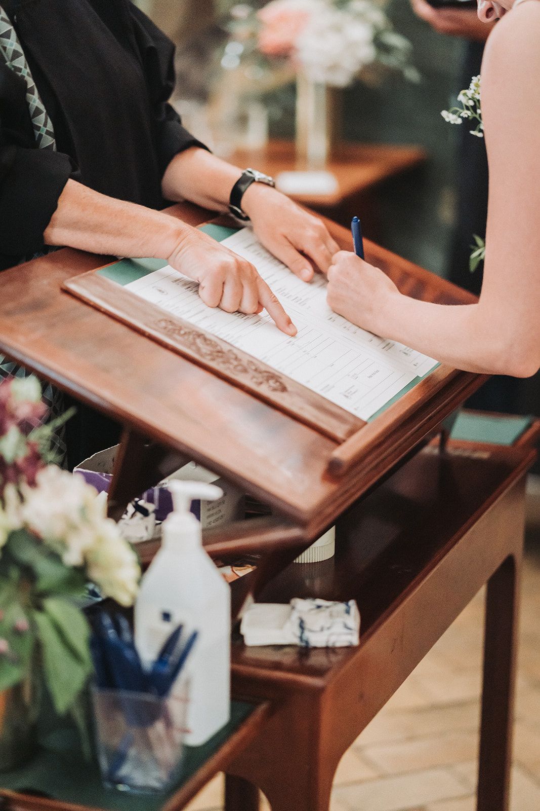 Hands sign an elegant wooden guest book at a formal event with floral decorations and a fountain pen.