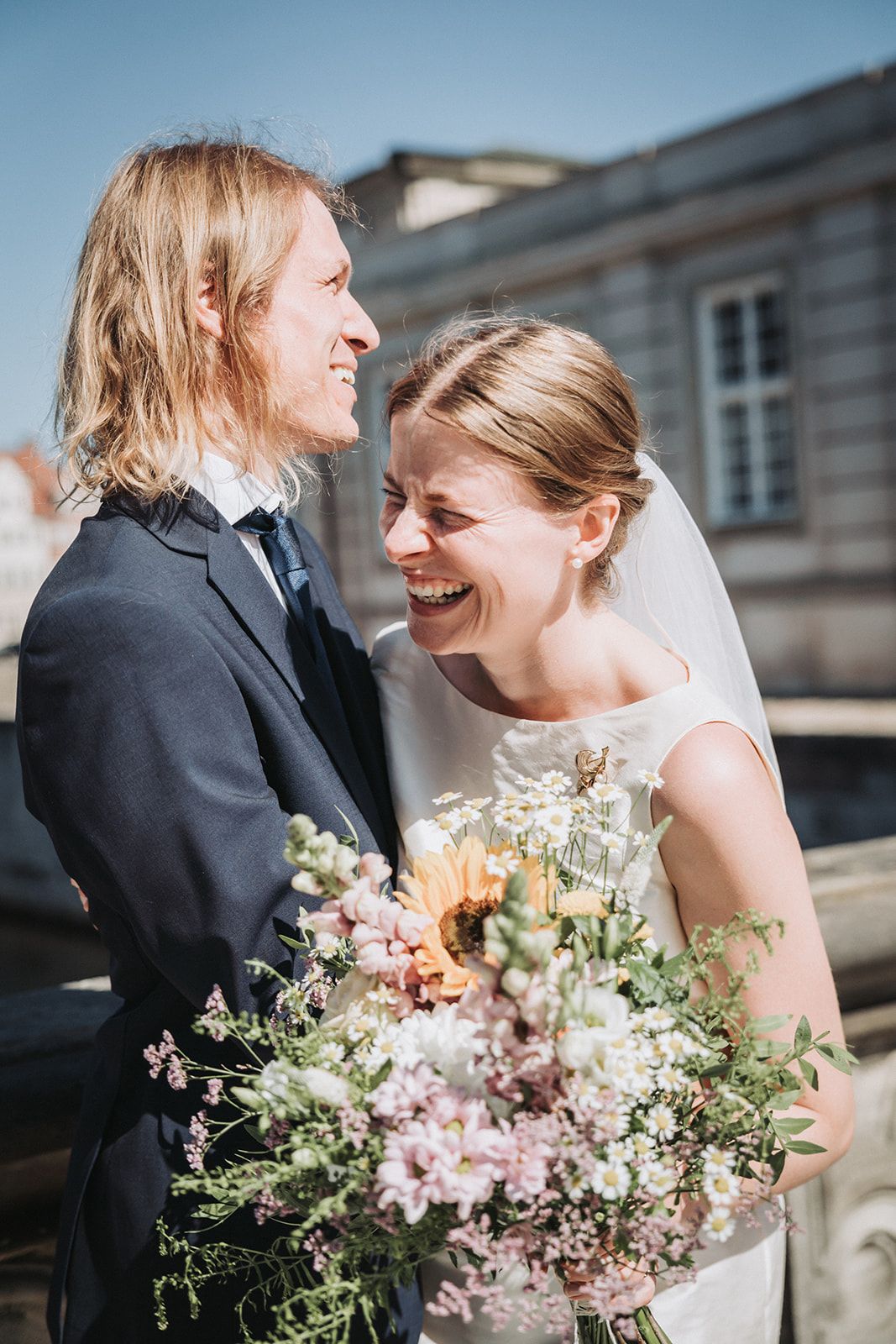 A joyful wedding couple shares a laugh while holding a pastel wildflower bouquet outdoors.