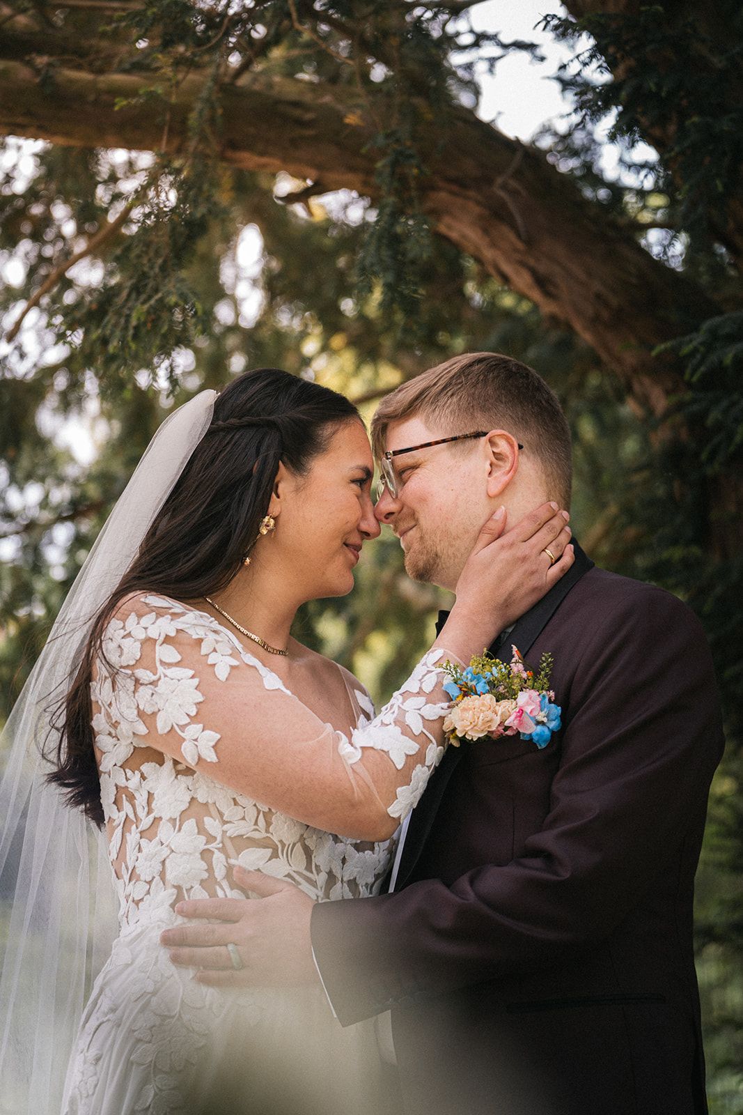 A tender romantic wedding moment captured outdoors under a natural tree archway with soft sunlight filtering through.