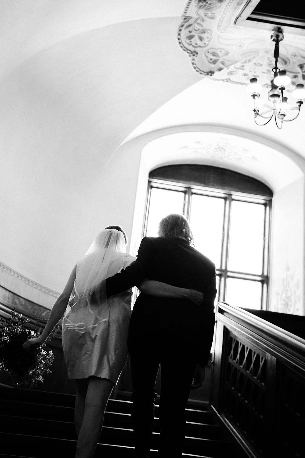 A black and white photo of bride and companion walking up elegant stairs with ornate chandelier and banister.