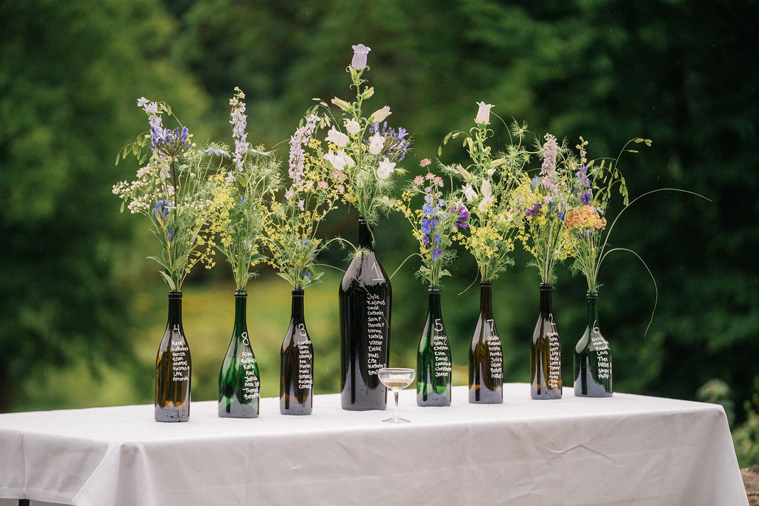 Wine and beer bottles repurposed as flower vases display wildflowers on a white tablecloth against a green backdrop.