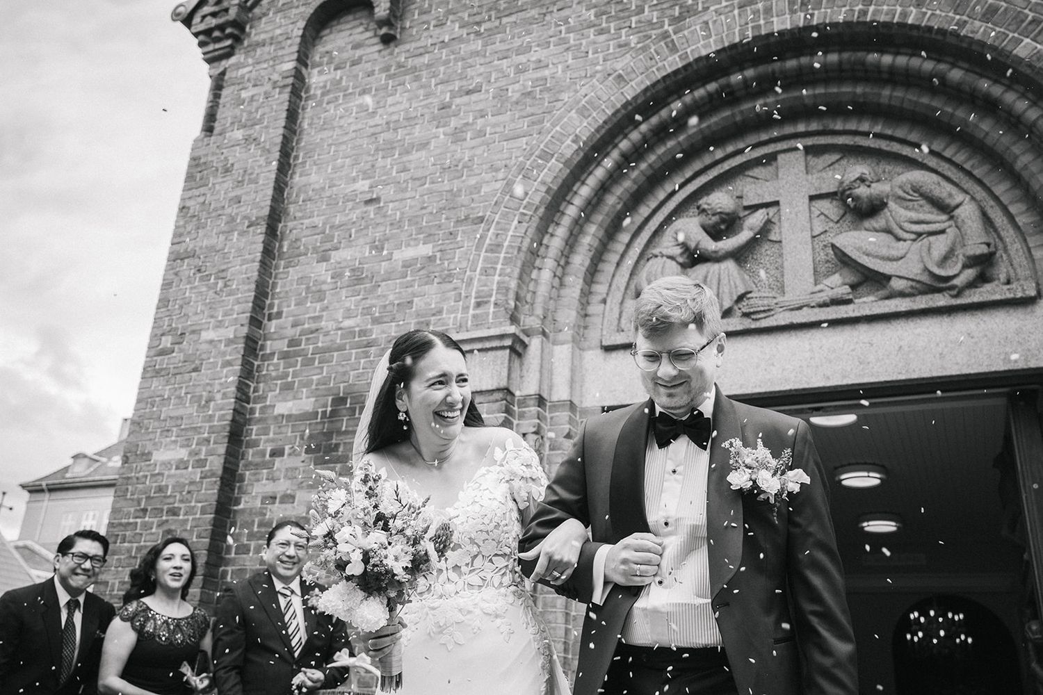 A happy couple exits a stone church after their wedding ceremony while guests celebrate by tossing confetti.