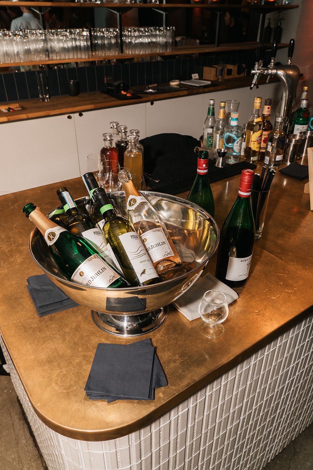 Silver ice bucket filled with chilled wine bottles on a wooden bar counter with napkins and glassware.