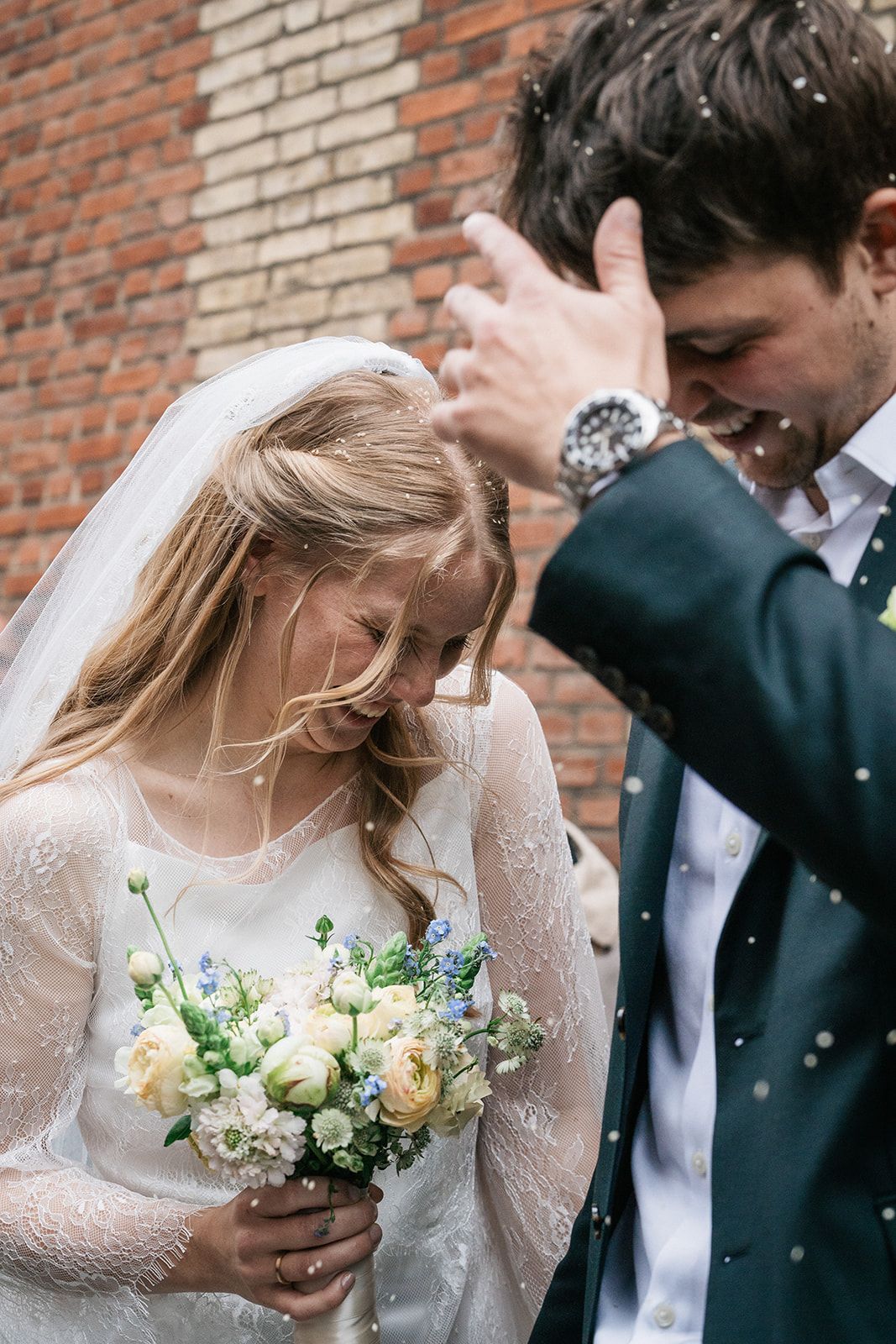 Happy newlyweds celebrate with confetti against a brick wall while bride holds a white and yellow floral bouquet.