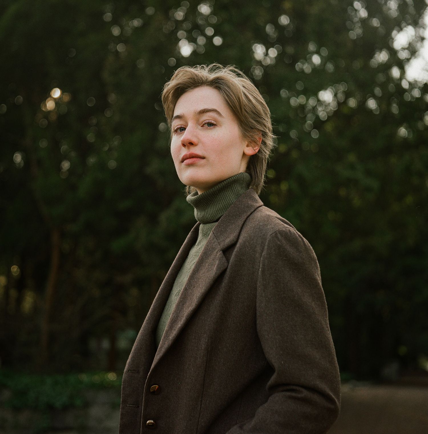 Person wearing brown coat and turtleneck poses outdoors with bokeh effect and trees in background during golden hour.
