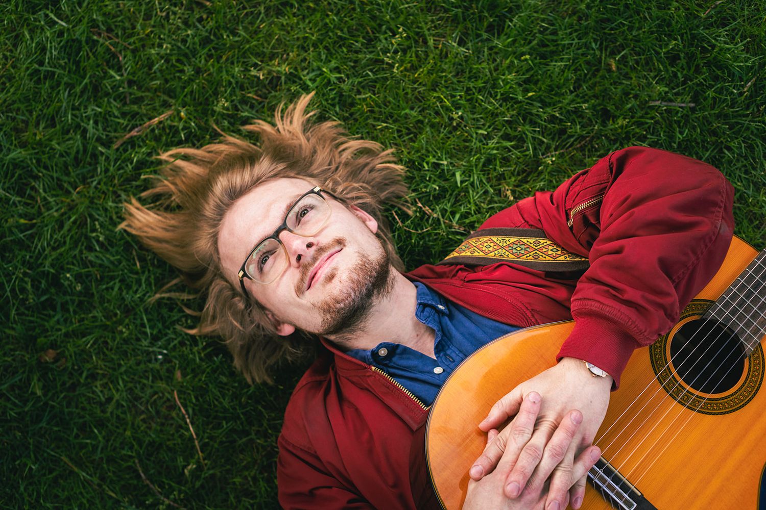 Musician in red jacket lying on grass with acoustic guitar while smiling at the camera.