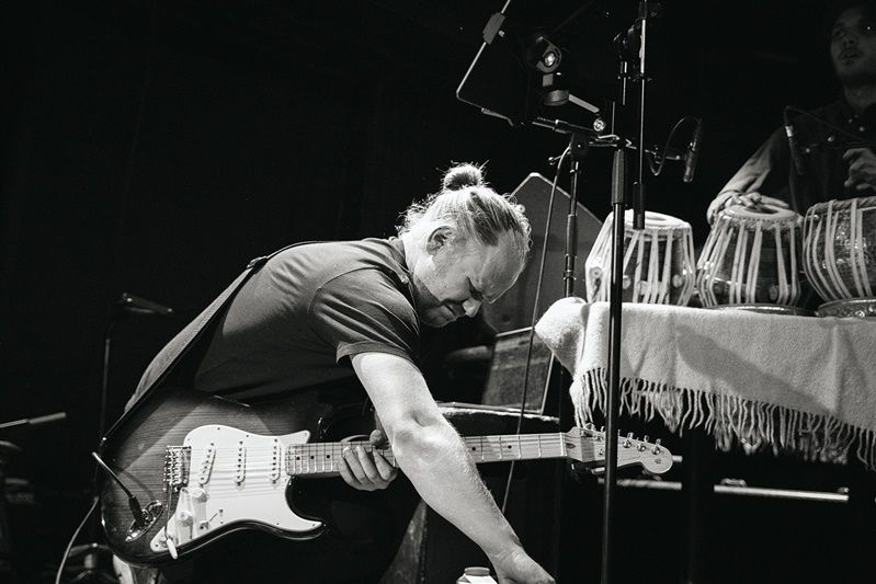 Black and white photo of an energetic guitarist bending down while performing on stage with a Stratocaster guitar.