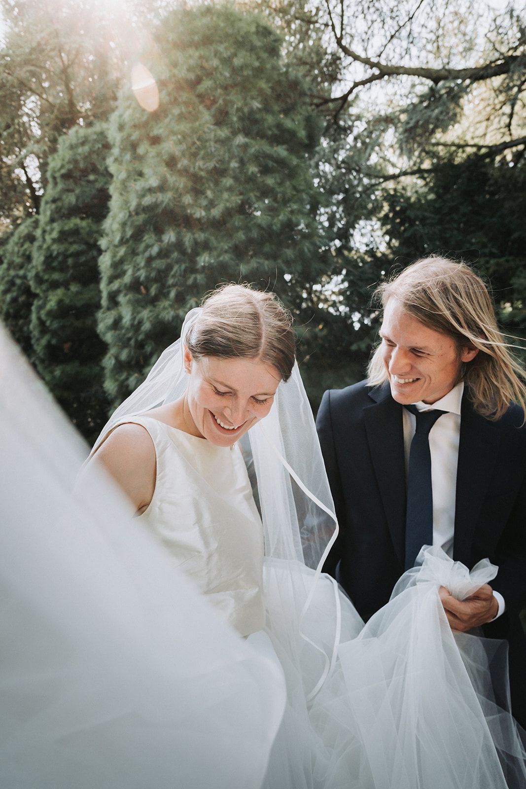 A happy wedding couple shares intimate laughter and loving moments in a wooded outdoor setting, surrounded by light and greenery.
