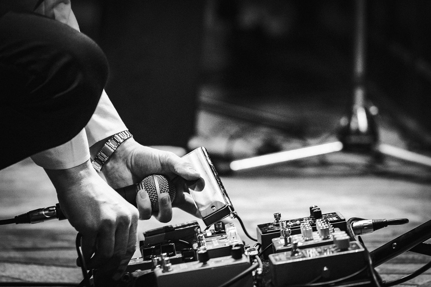 A close-up black and white shot of hands working with a guitar pedal or effects unit on a wooden surface.