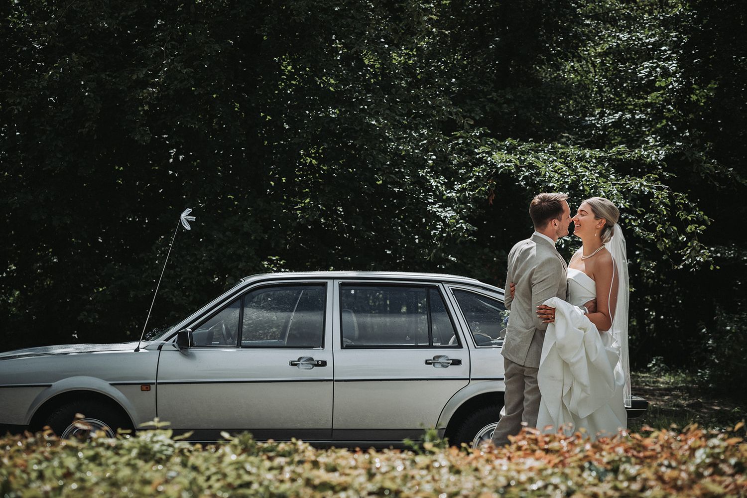 A silver vintage car is parked in a wooded area with fallen autumn leaves while a couple shares a tender moment.